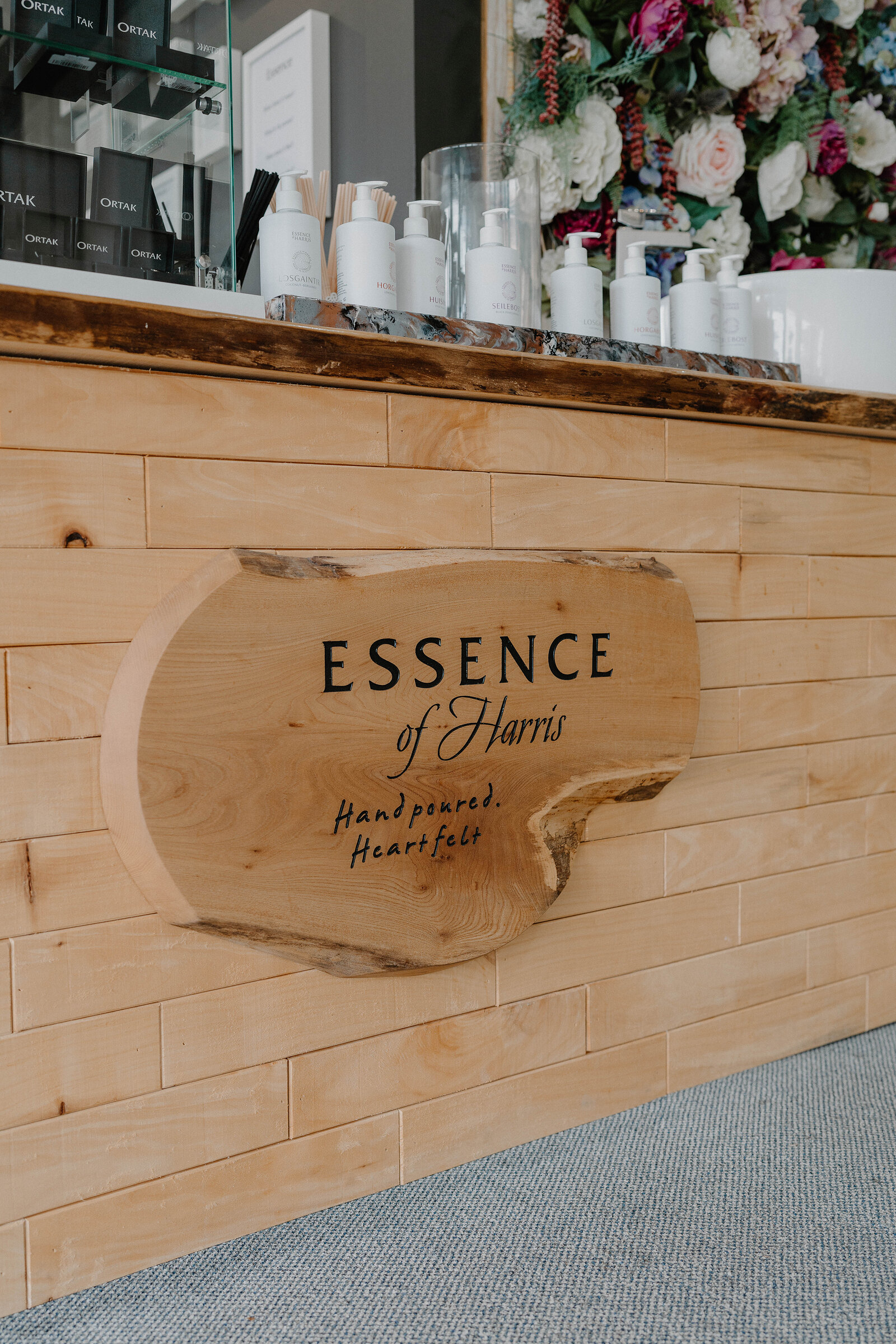 A carved wooden sign reading Essence of Harris, Hand poured, Heartfelt, set into a wood-panelled shop counter, with pump-top bottles and a floral display on the shelf above.