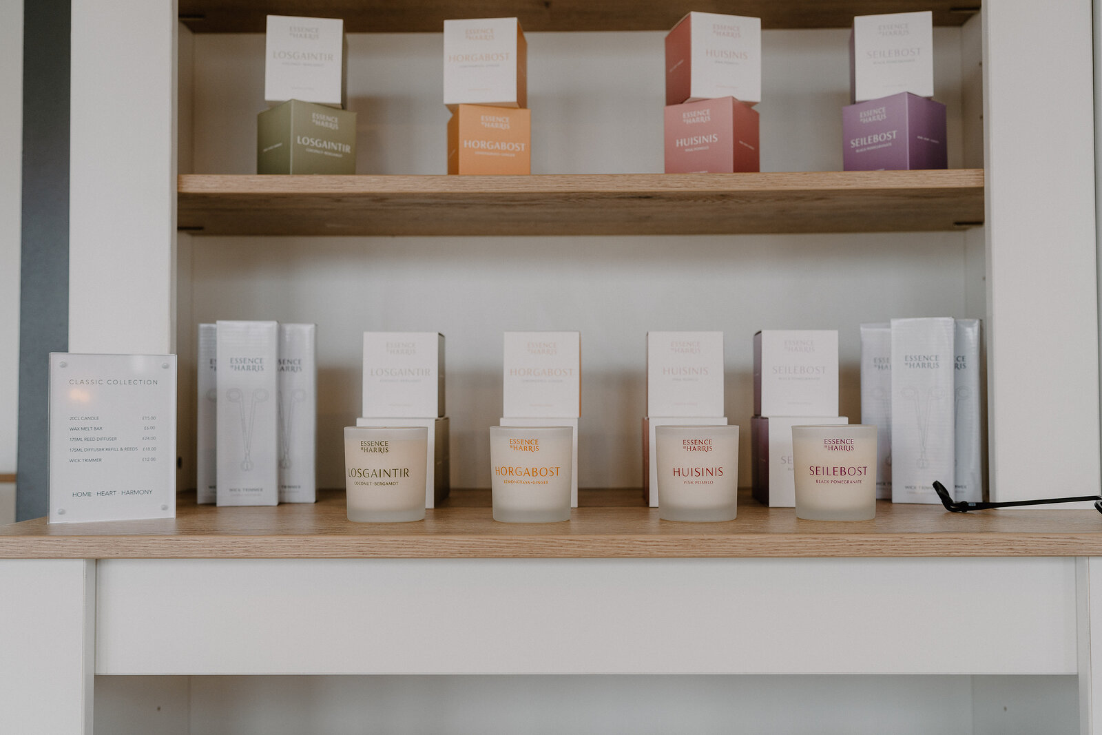 A retail shelf holding four labelled candles in frosted glass jars, including Bergamot, Verbena and Wildrose, with matching gift boxes on the shelf above and a folded price list to the left.
