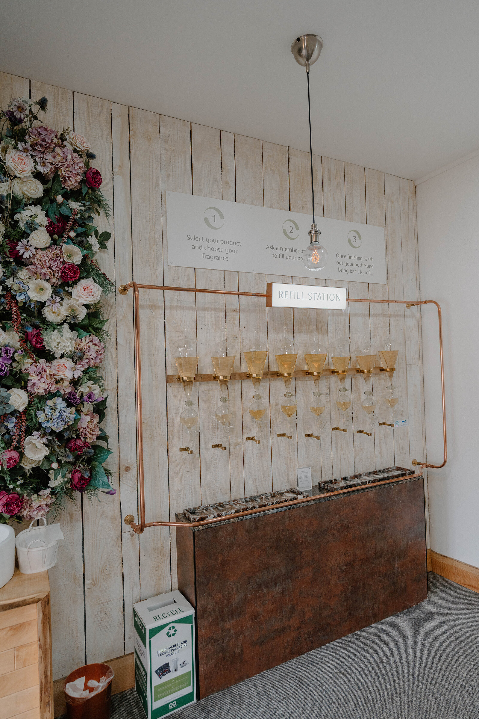 A copper-piped refill station holds a row of glass bottles filling with pale liquid beneath a sign reading Refill Station, a wall of artificial flowers to one side and a recycling bin below.