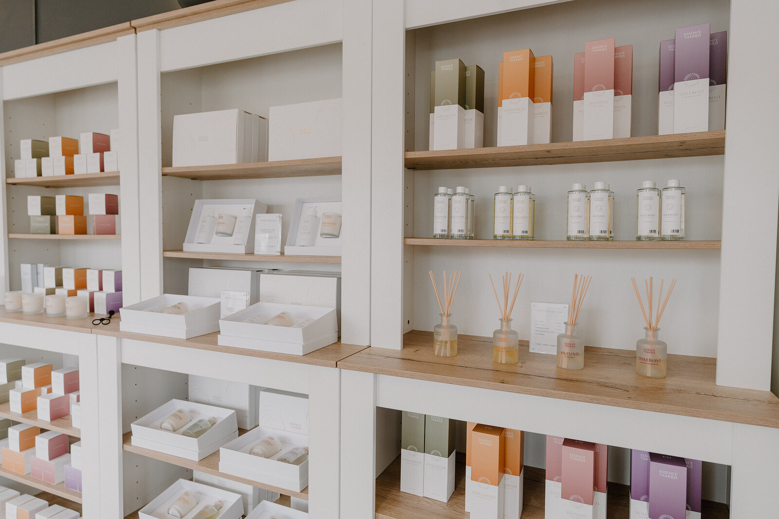 A shop shelf of skincare boxes and pump bottles arranged by colour, with a row of reed diffusers on the lower shelf.