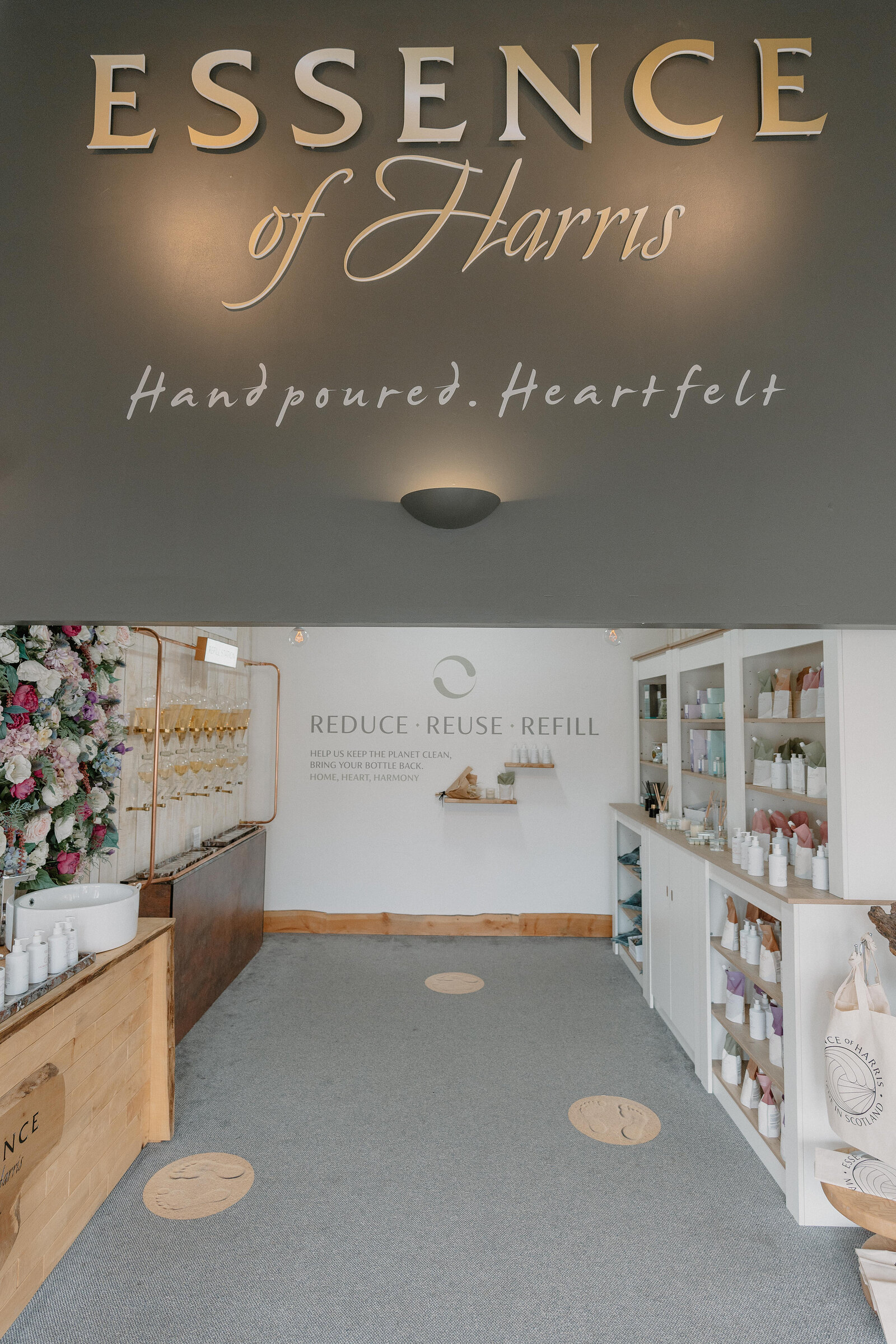 Gold lettering reading Essence of Harris, Hand poured, Heartfelt, hangs above a corridor leading to a refill station sign urging Reduce, Reuse, Refill, shop shelving and a floral display either side.