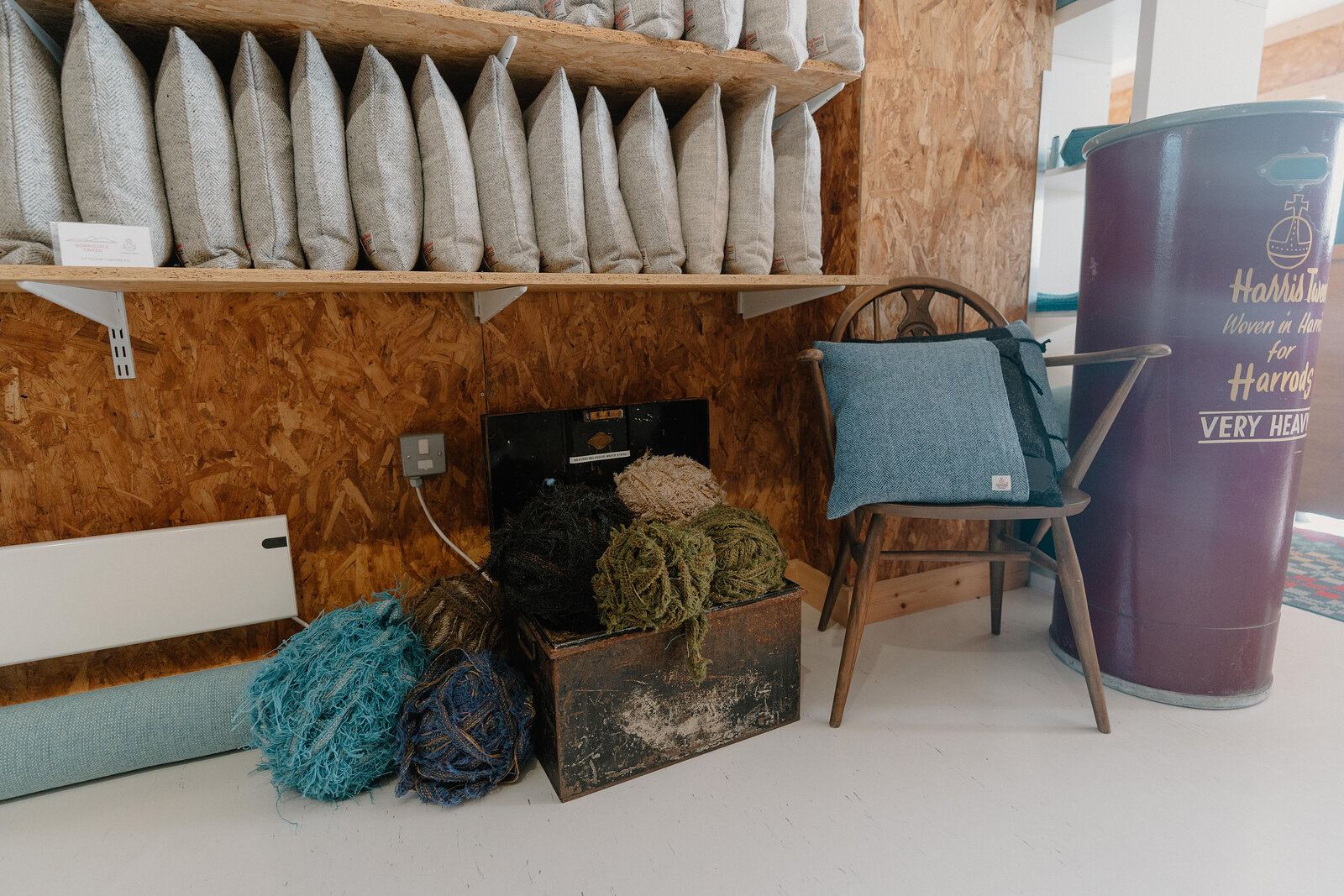 Rolled bolts of tweed fabric fill a wooden shelf above a crate of coloured yarn skeins, a chair with a tweed cushion and a vintage advertising tin standing nearby.