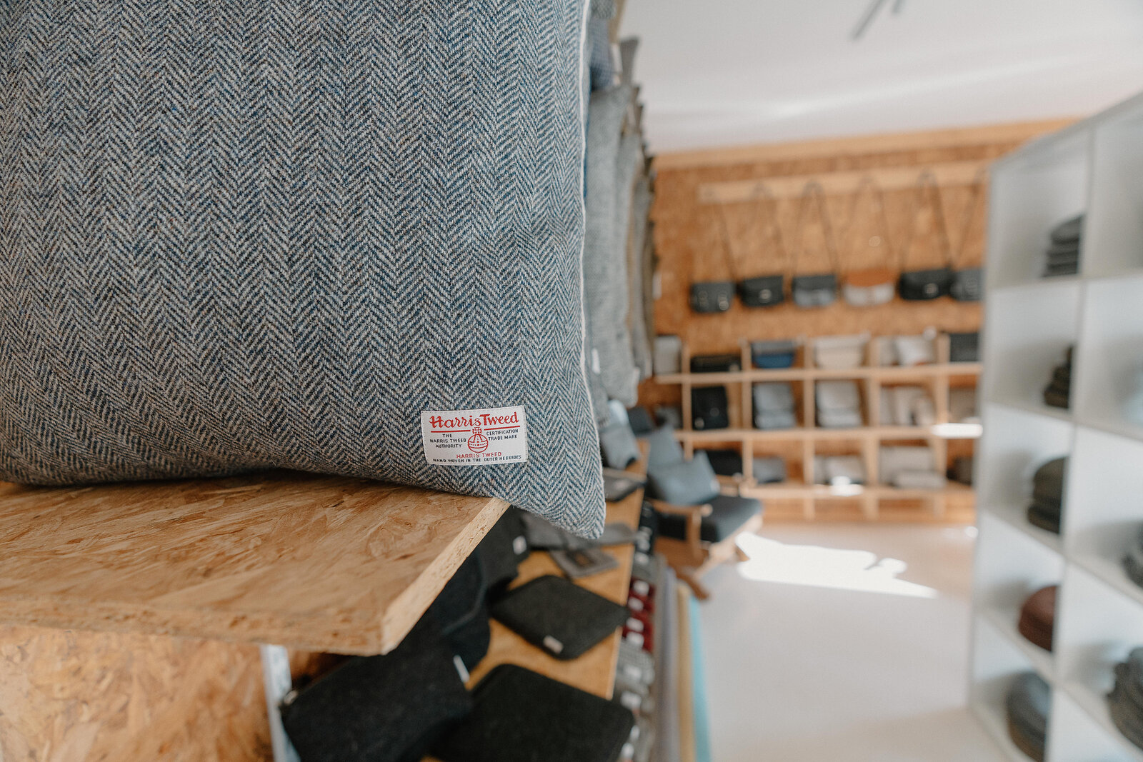 A grey herringbone tweed cushion labelled "Harris Tweed" rests on a wooden shelf, rows of fabric samples and folded cloth visible behind.
