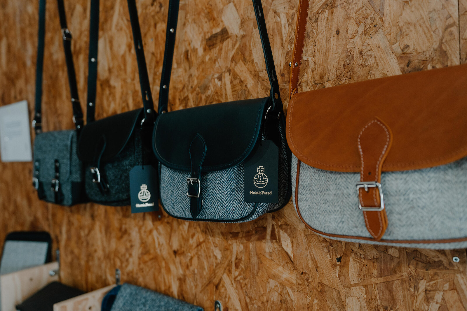 Tweed satchel bags with leather trim hang in a row from pegs on a wooden board, a brand tag visible on the strap.