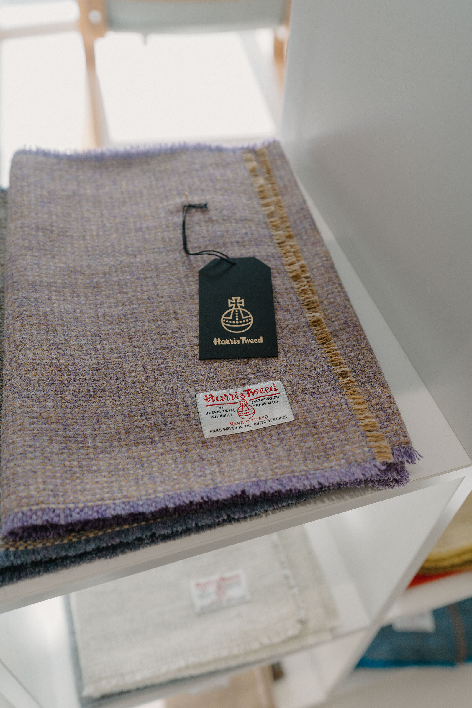 A folded length of purple and gold-flecked Harris Tweed fabric lies on a shelf, its brand tag and label visible.