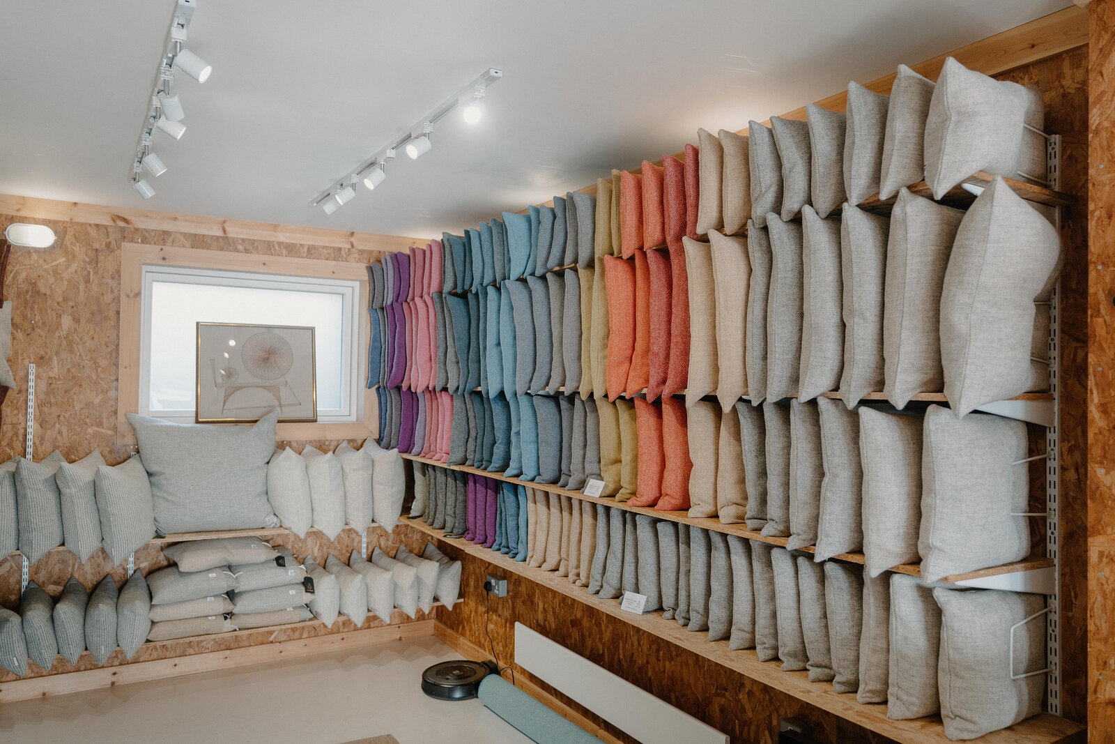 Rows of folded tweed fabric in varied colours fill tall wooden shelving on both sides of a narrow shop, a bench of cushions at the far end.