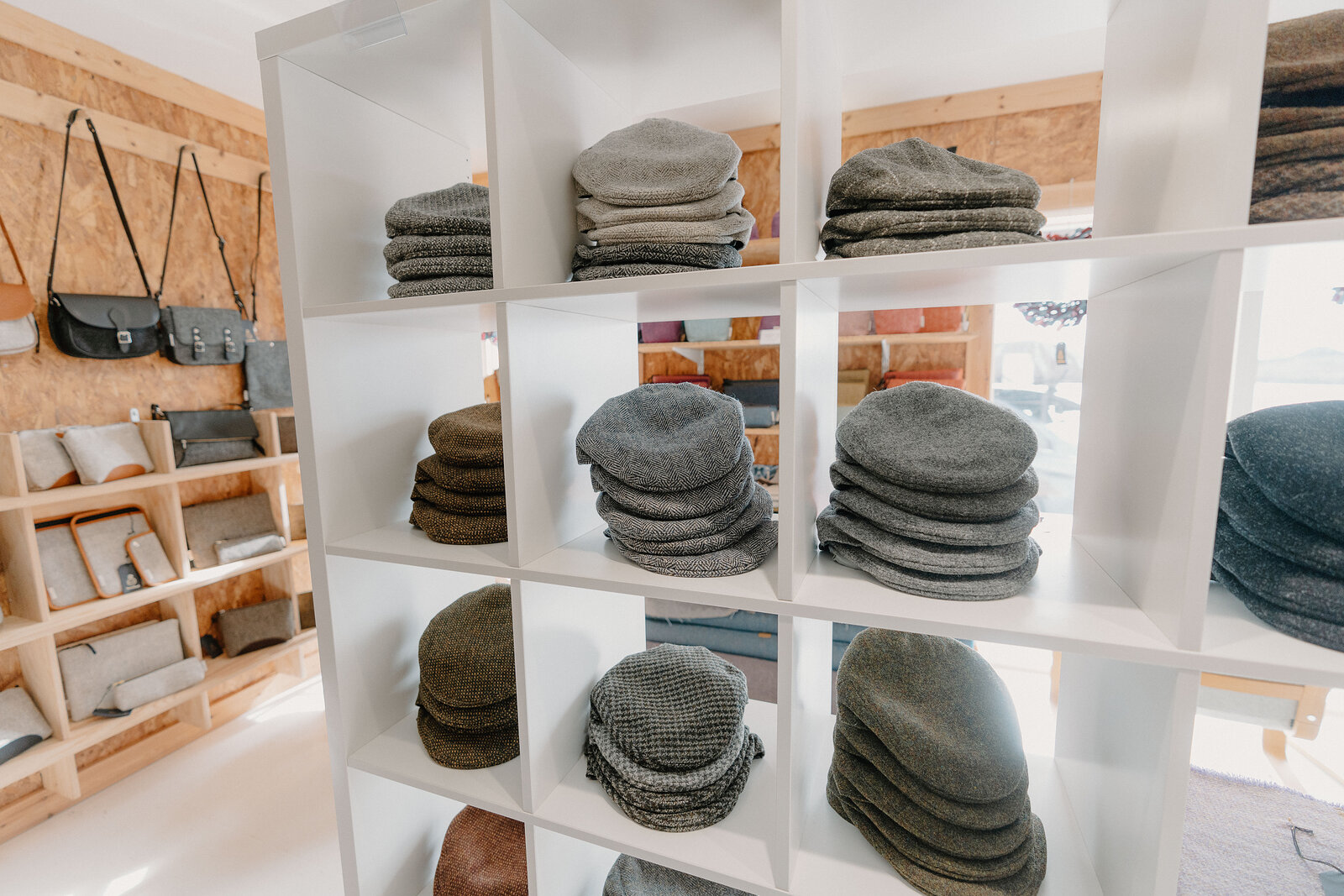 Flat caps and tweed hats are stacked on white cube shelving beside handbags and folded scarves.
