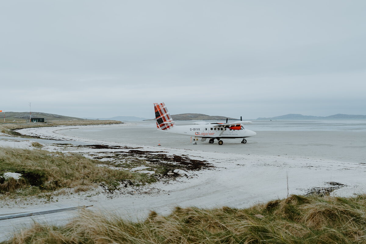 A small propeller aircraft with a red-and-white checked tailfin standing on a wet tidal beach runway, low dunes and marram grass in the foreground.
