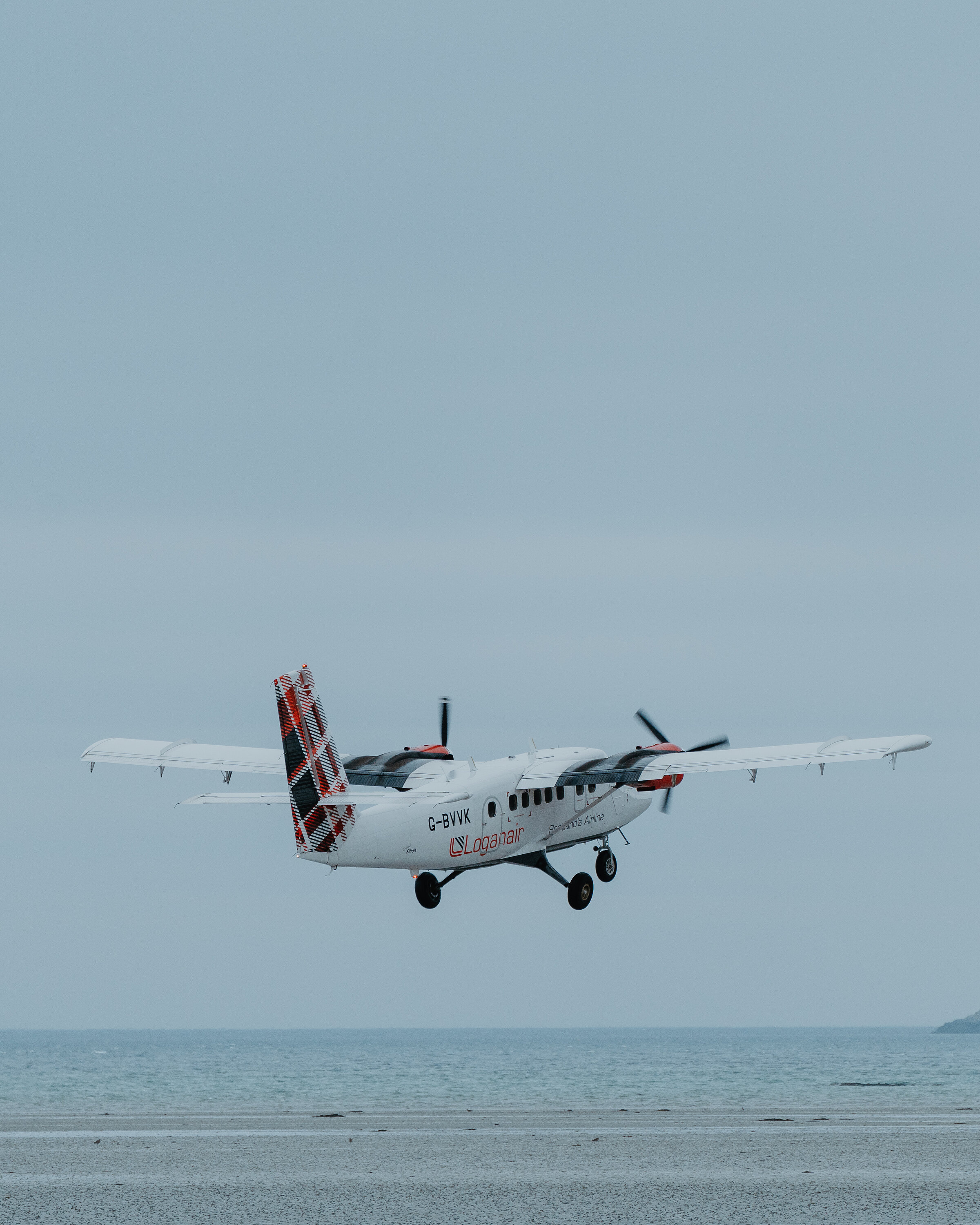 A twin-propeller aircraft comes in to land low over the shore, its tartan tailfin bright against a pale sky and landing gear extended above the sand.