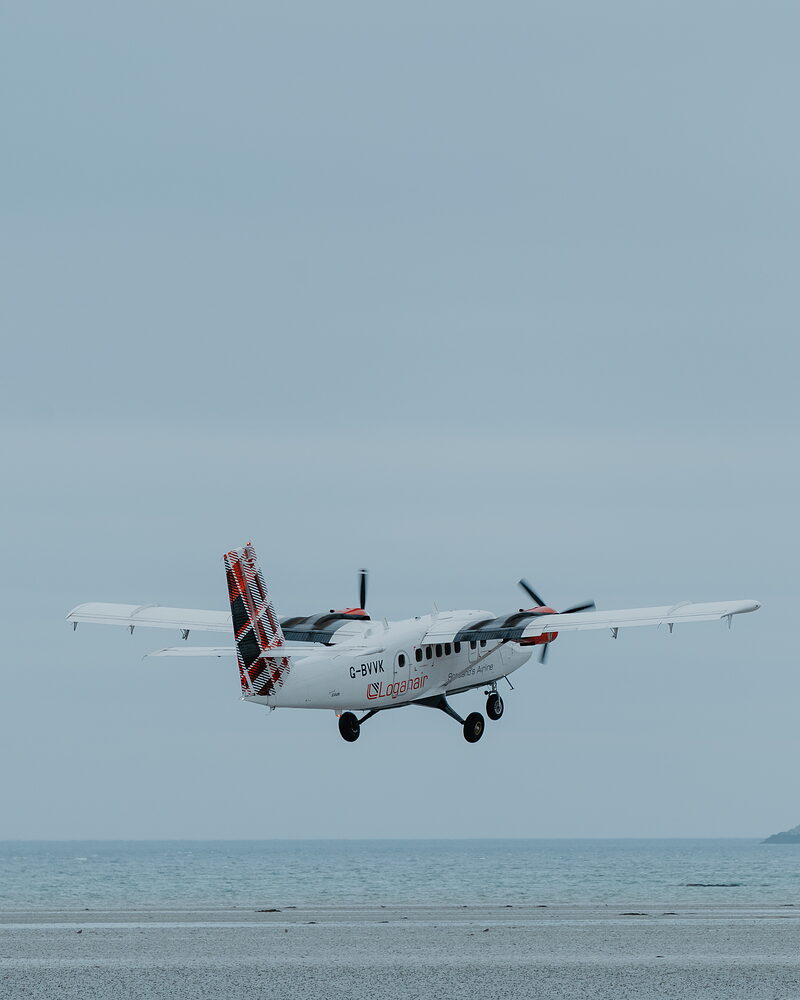 A twin-propeller aircraft comes in to land low over the shore, its tartan tailfin bright against a pale sky and landing gear extended above the sand.