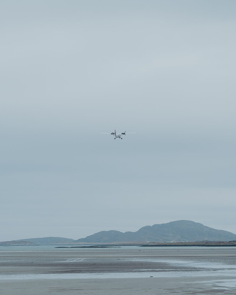 A small aircraft flies low over a wide tidal beach, a hill rising behind and the sand stretching pale beneath a grey sky.