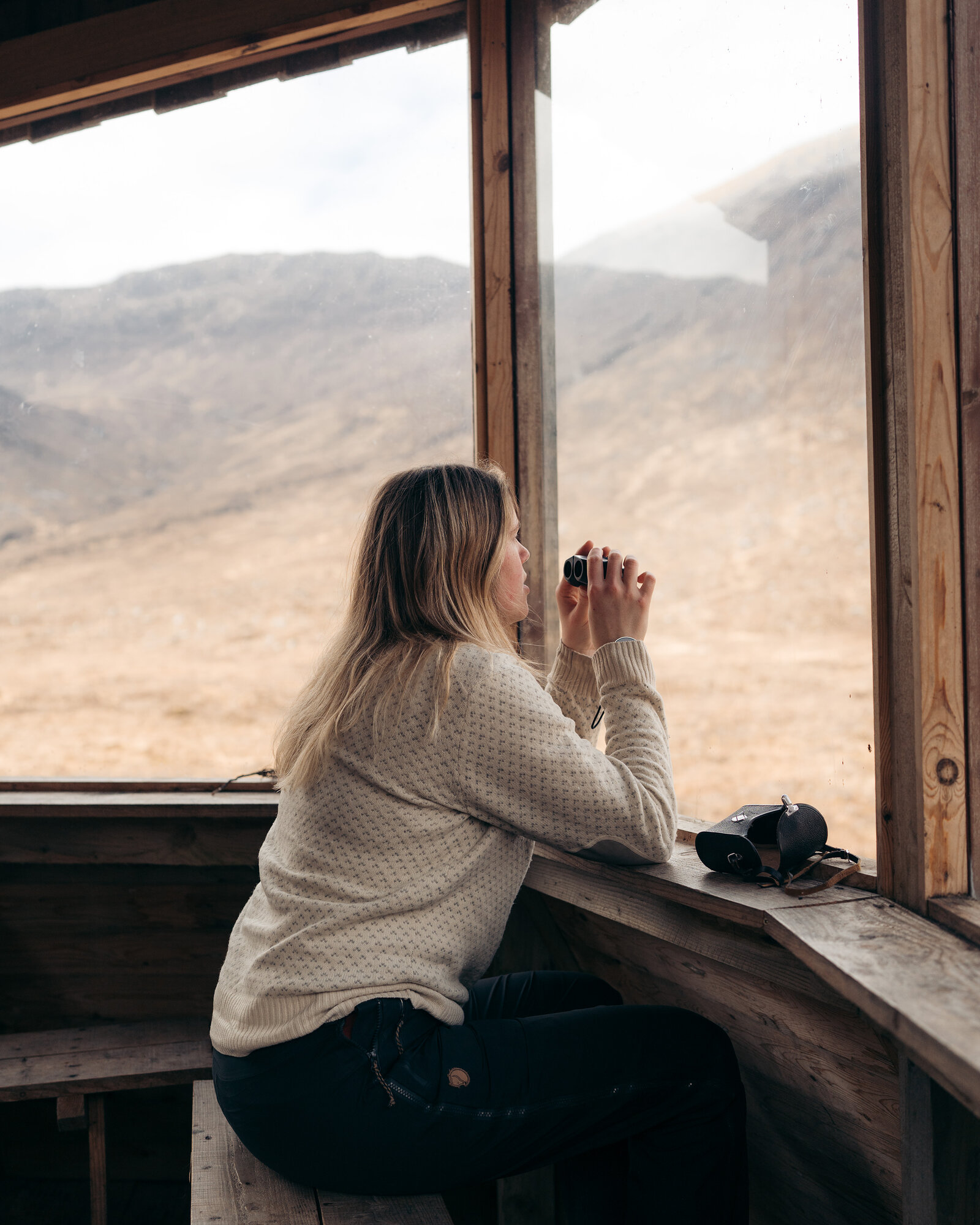 A woman looks through binoculars from a wooden hide window overlooking a misty mountain landscape.