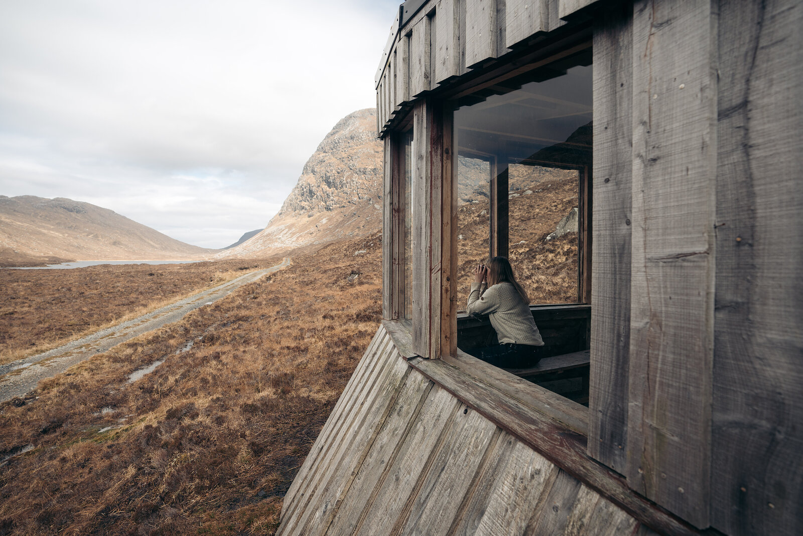 A person sits inside a wooden hide overlooking a mountain glen, a road and river visible through the open window.