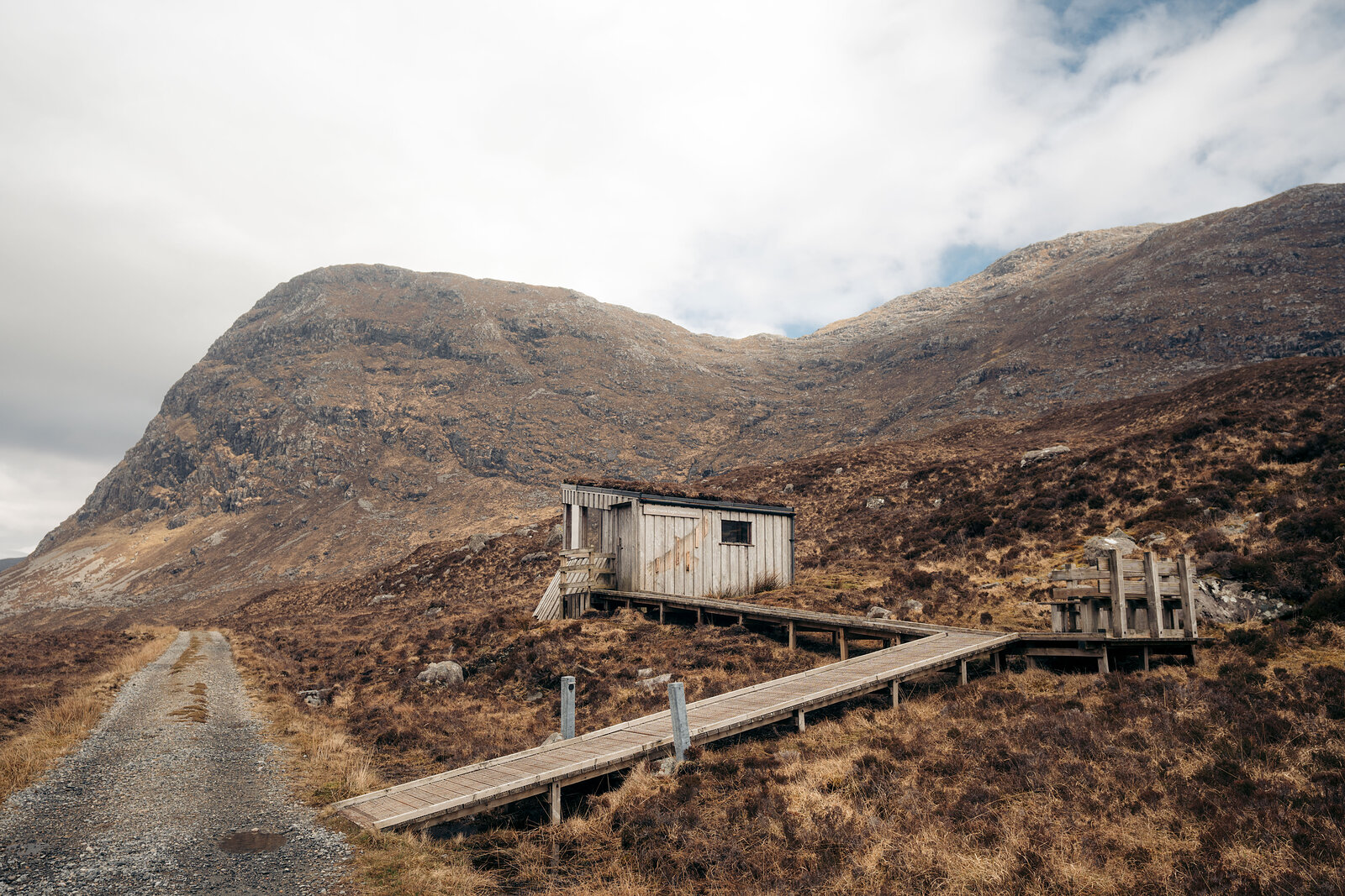 A weathered timber hut stands beside a boardwalk and gravel track, a large rounded mountain rising behind it.