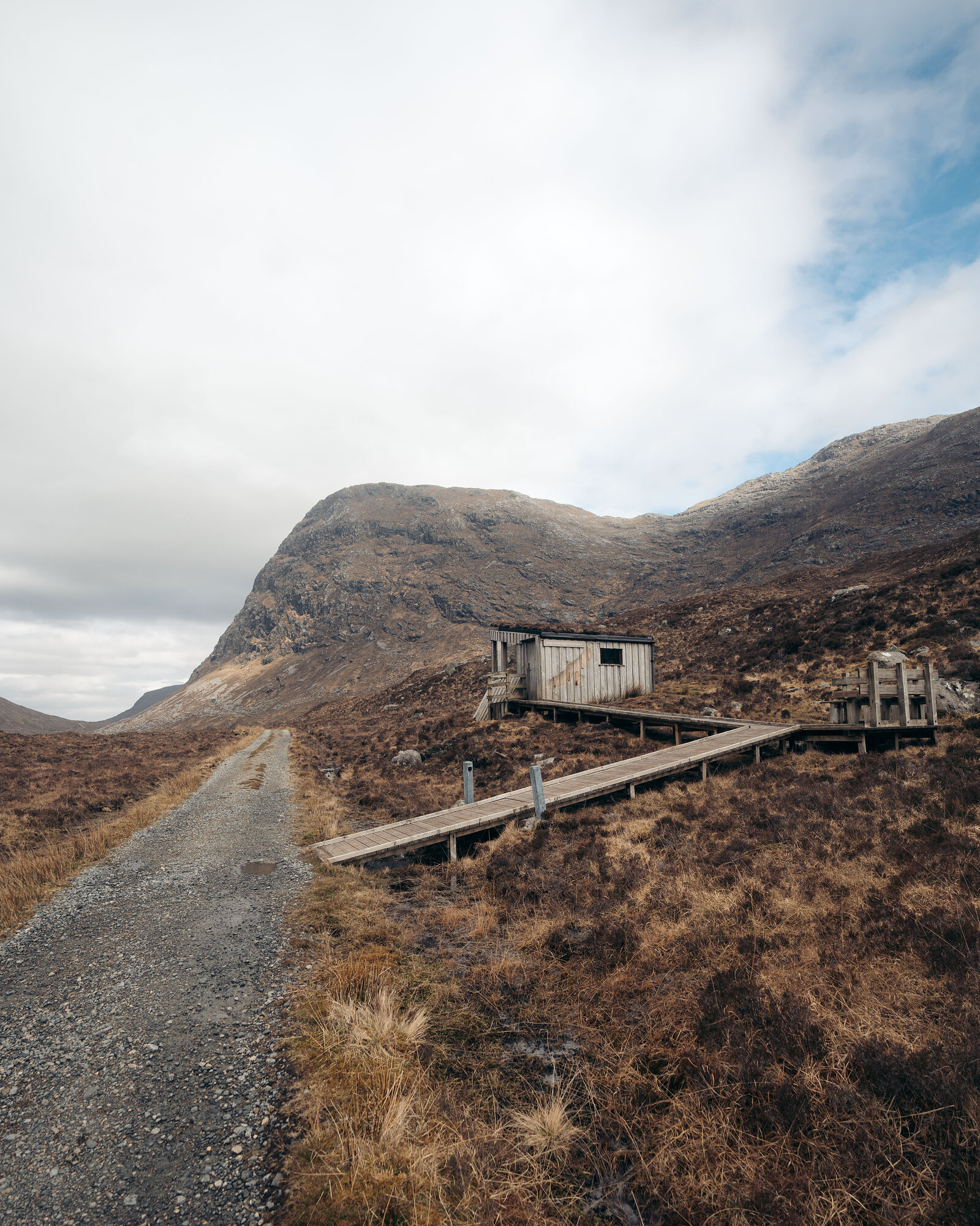 A weathered timber hut stands beside a boardwalk and gravel track, a large rounded mountain rising behind it.