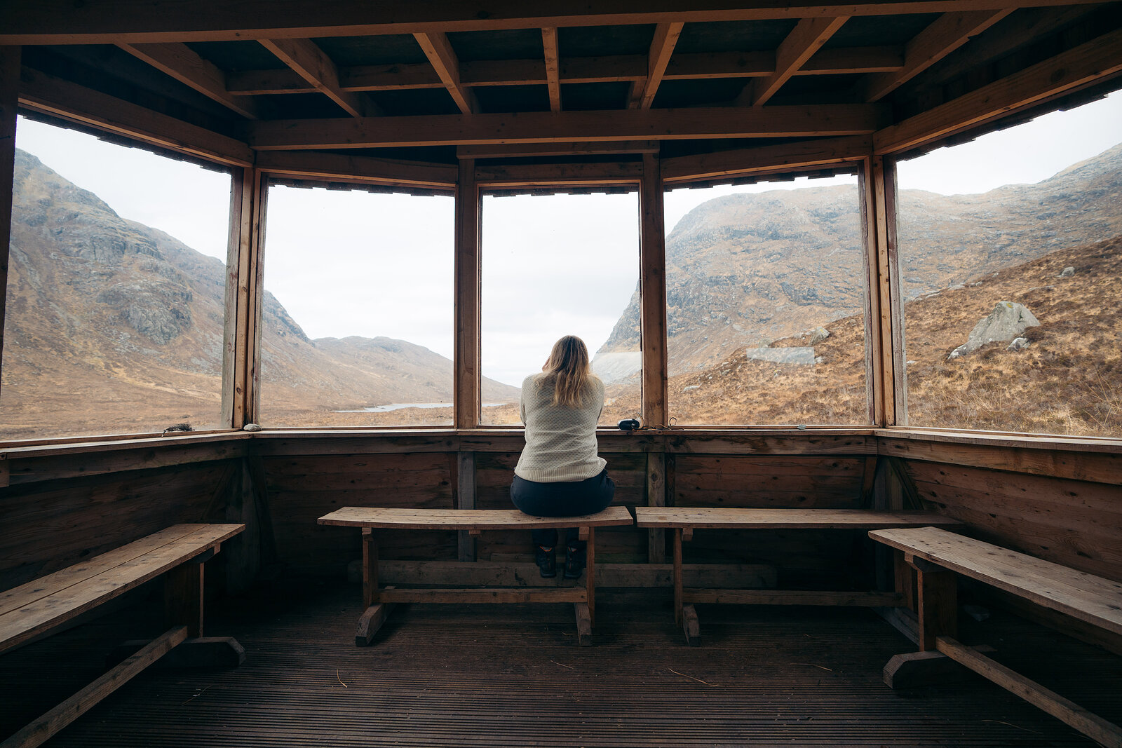 A woman sits alone on a wooden bench inside a hexagonal timber hide, looking out through large windows at a misty mountain glen.