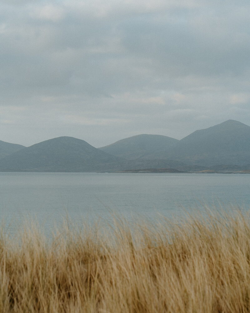 A calm sea loch with layered hills rising beyond the far shore, and a foreground of golden dune grass.