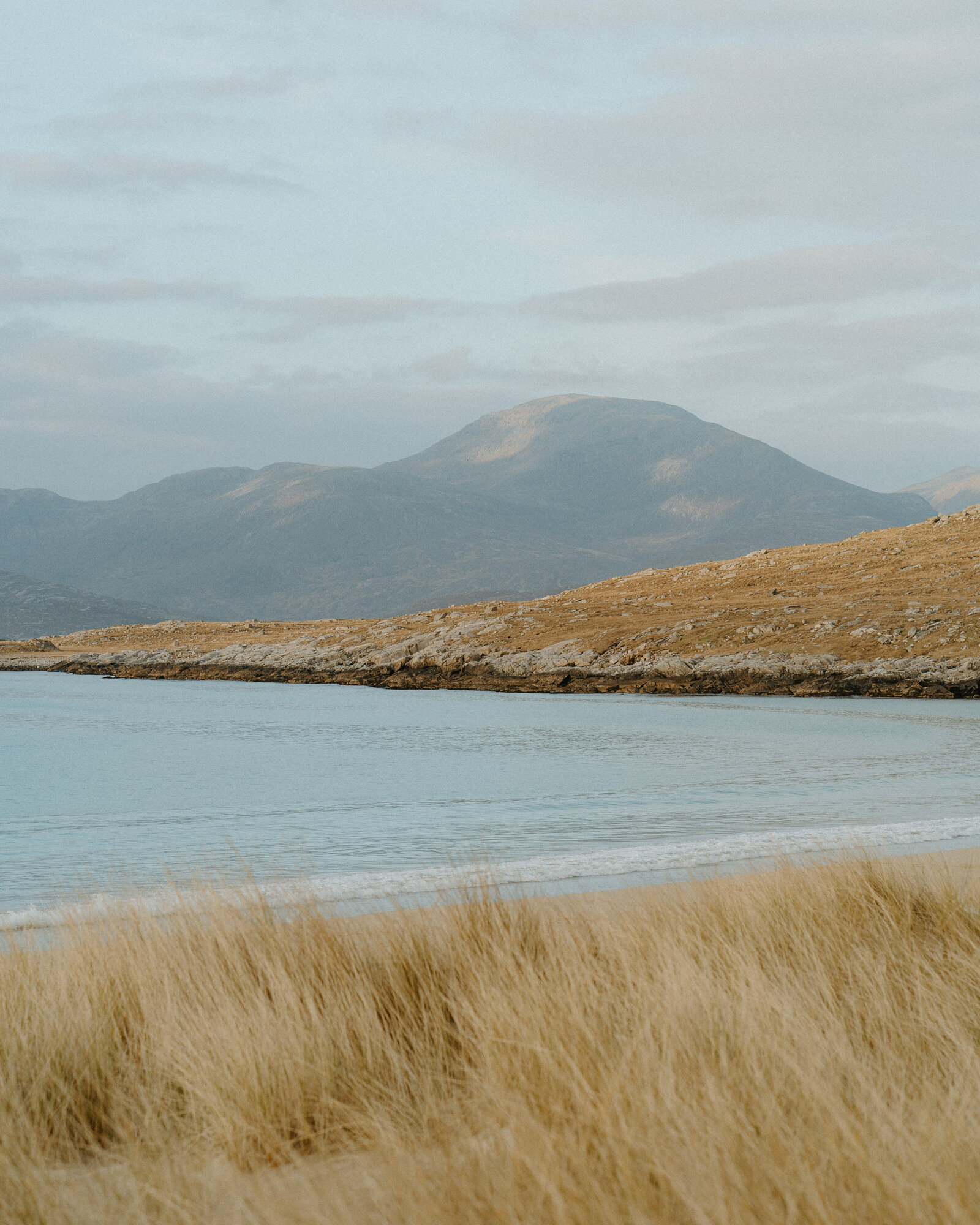 A single hill rising beyond a calm sea loch, seen across a shoreline of rock and grass with golden dune grass in the foreground.