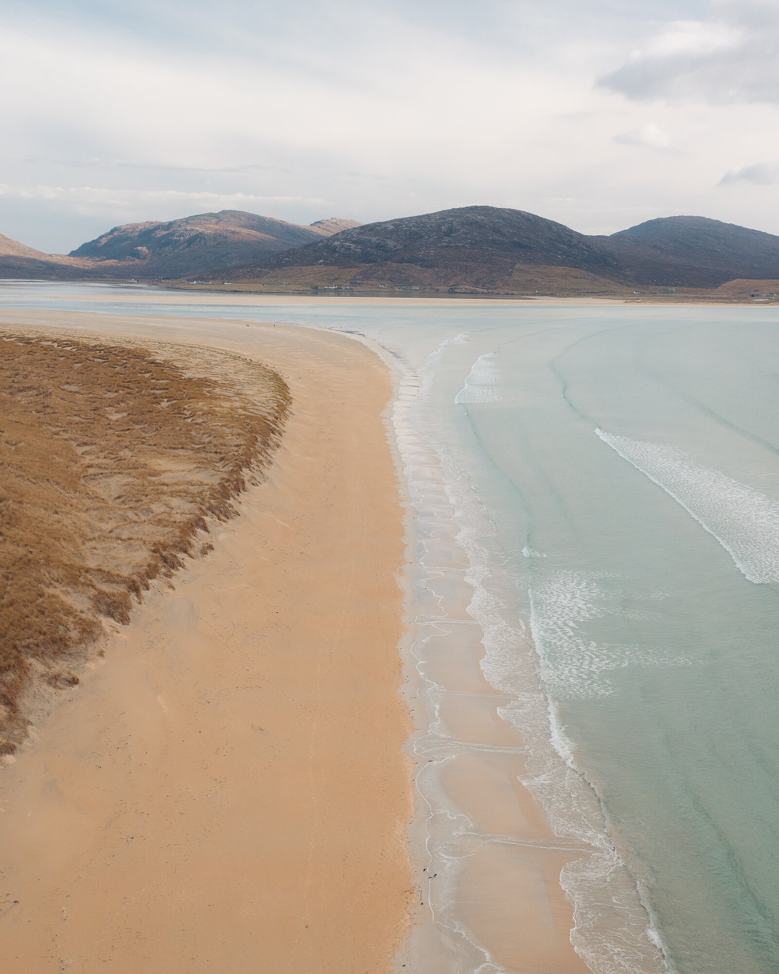 An aerial view of a long straight beach where pale turquoise waves break against golden sand, dune grass to one side and hills rising beyond a calm bay.