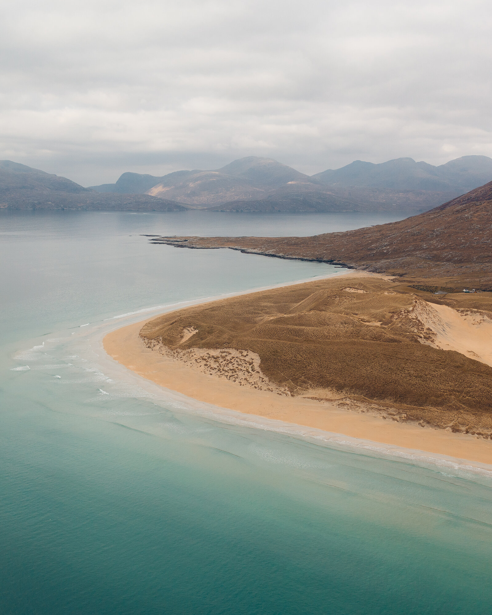 An aerial view of a curved sand spit reaching into turquoise water, dune grass covering its spine and hills rising across the bay beyond.