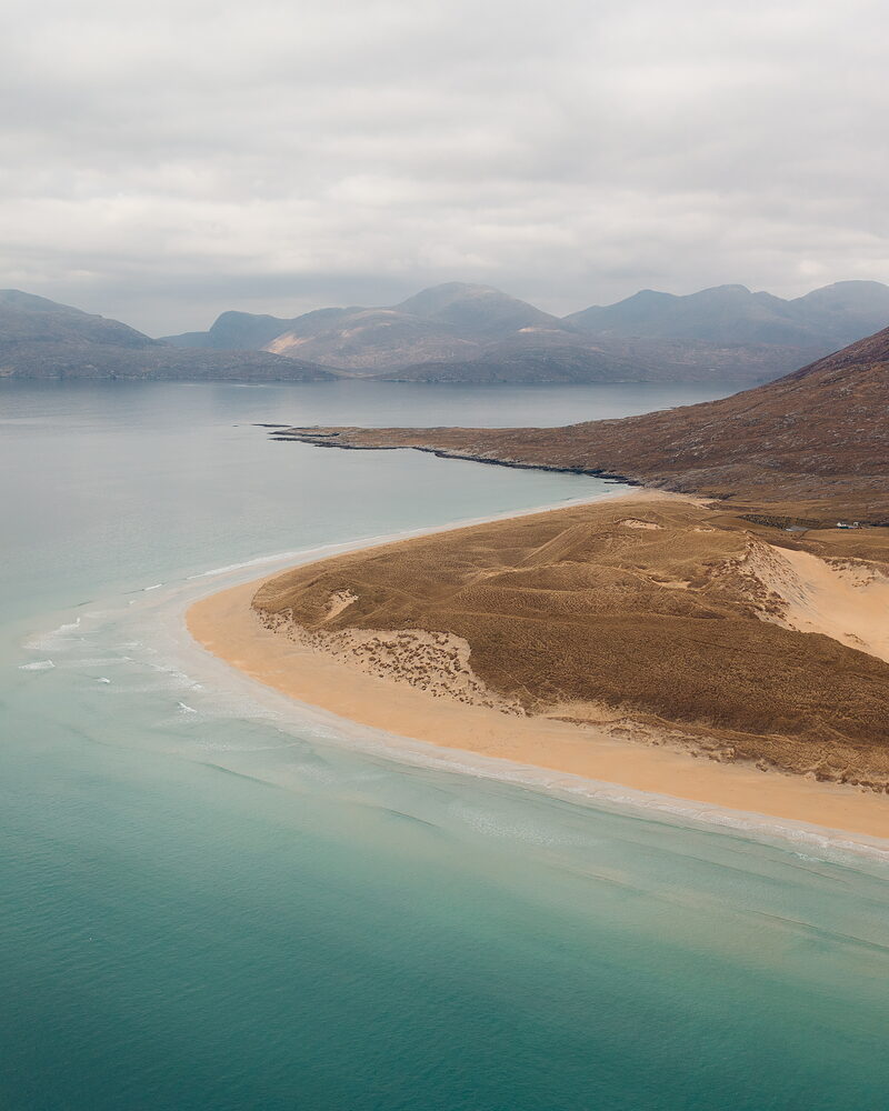An aerial view of a curved sand spit reaching into turquoise water, dune grass covering its spine and hills rising across the bay beyond.