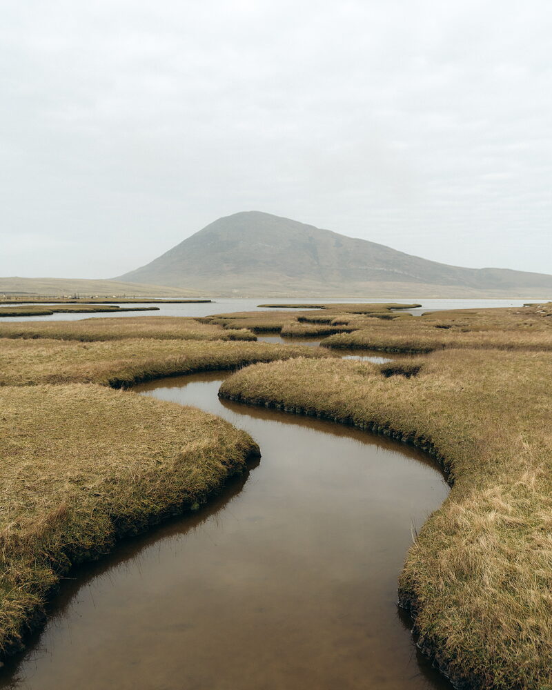 A tidal channel winding through golden marsh grass towards a still loch, with a single conical hill rising behind.