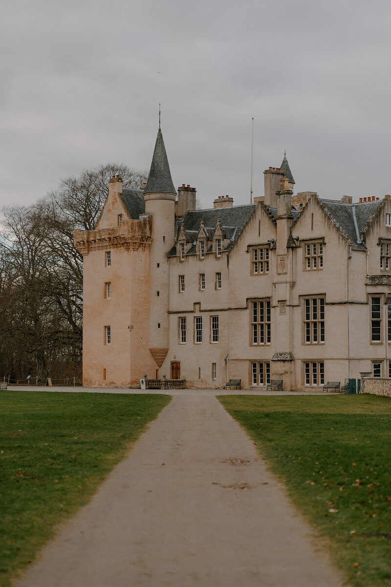 A turreted stone castle with crow-stepped gables stands at the end of a gravel path, bare trees to one side and mown lawns stretching away from its facade.