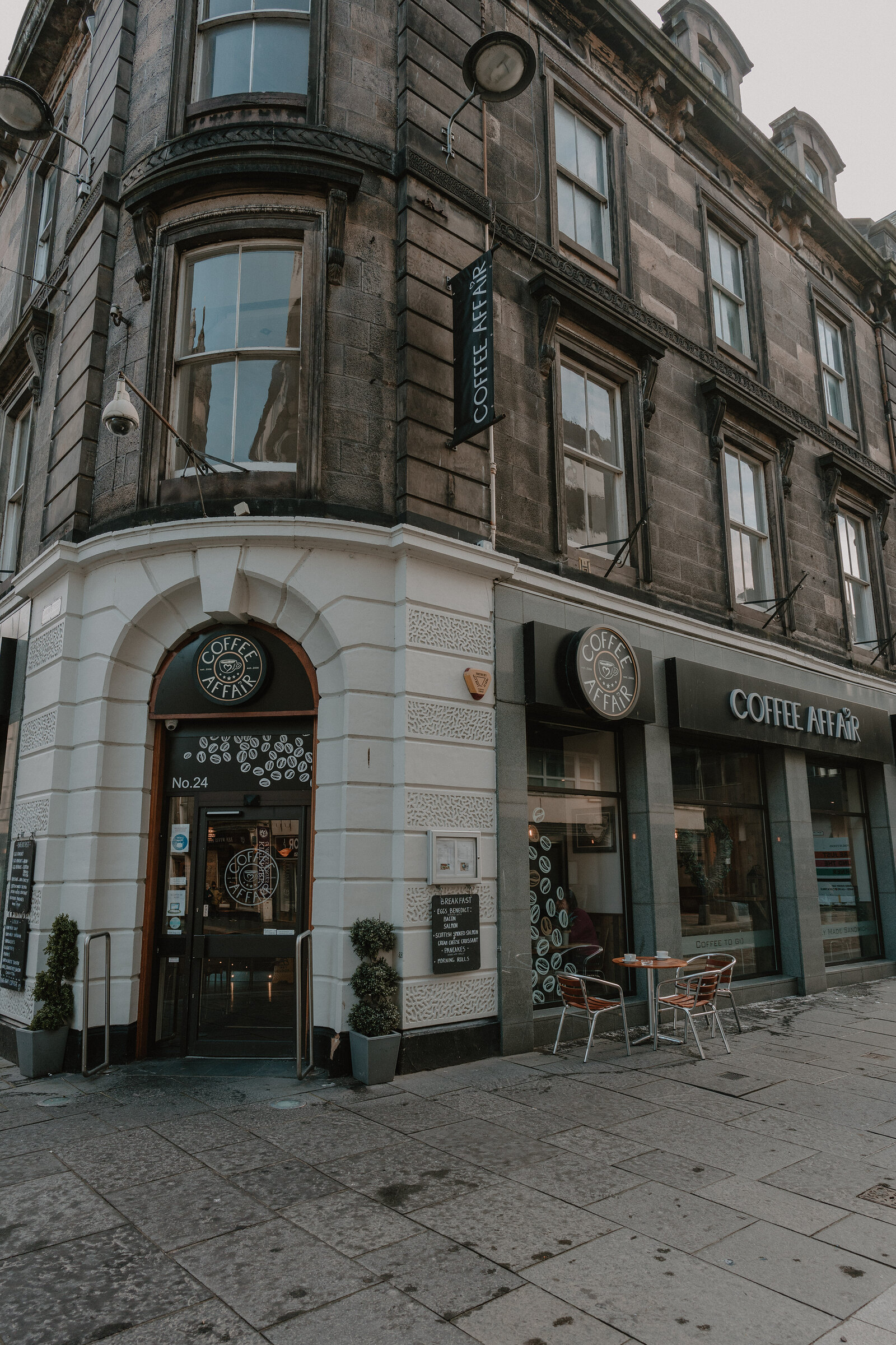 A curved corner shopfront numbered 24 is lettered "Coffee at No.1" and "Coffee Affair", outdoor tables set on the pavement beside potted plants.
