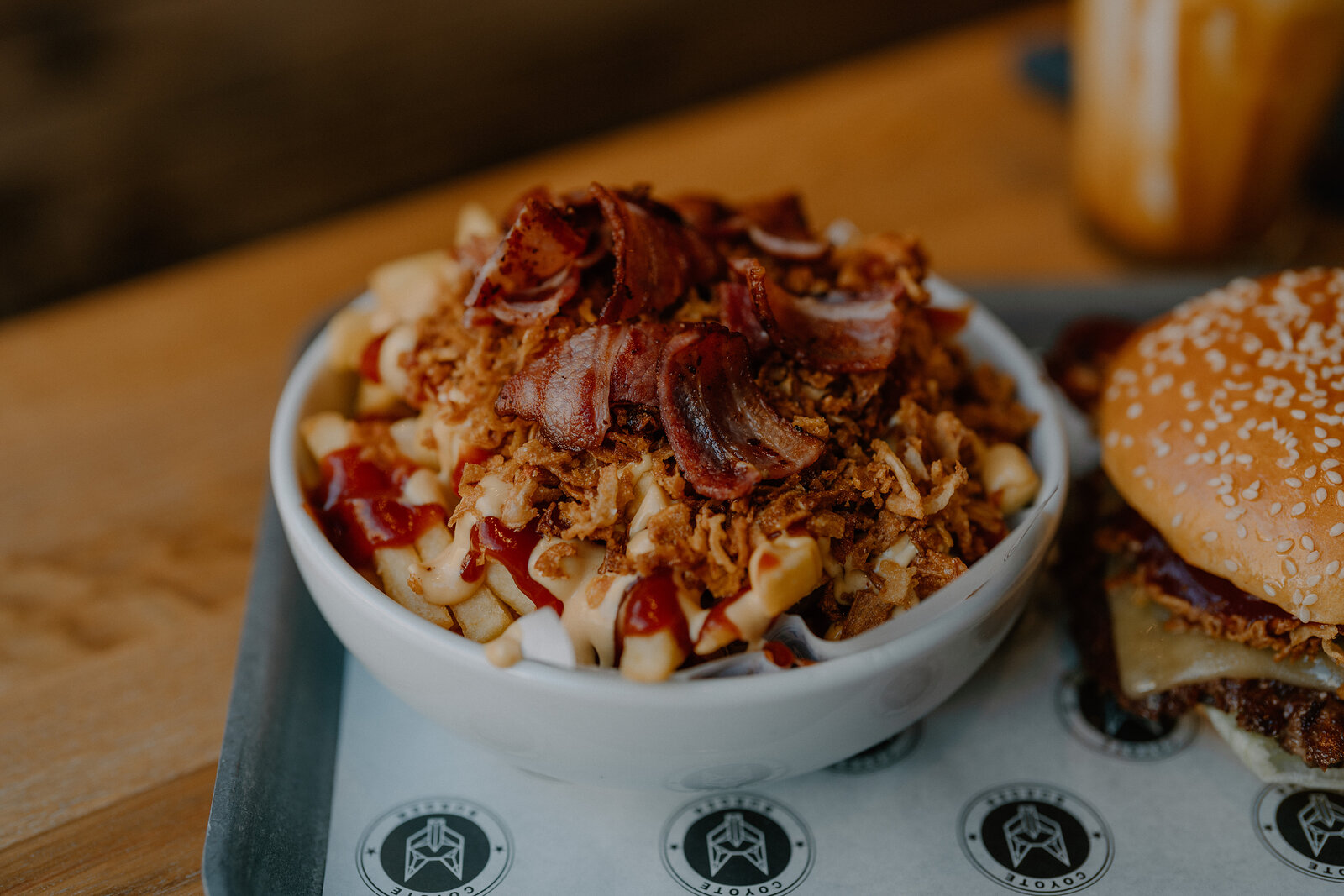 A bowl of loaded fries topped with bacon and crispy onions sits on a tray beside a sesame burger, a drink blurred behind.