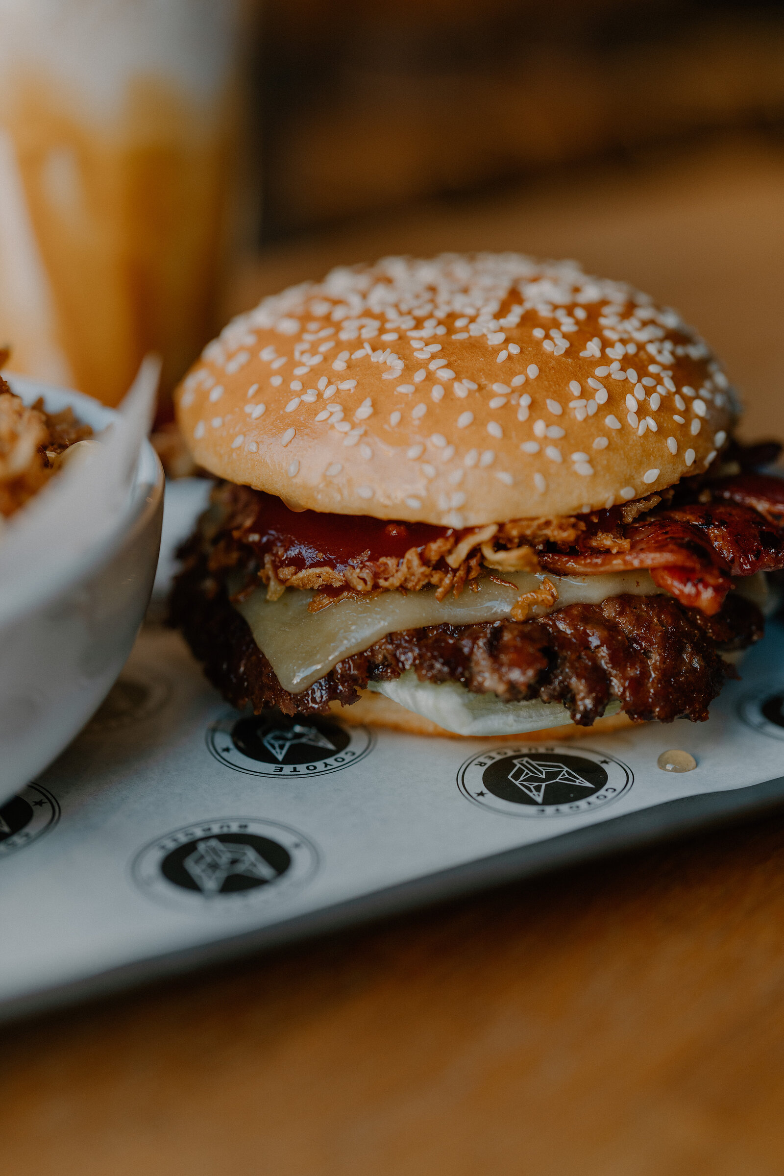 A sesame bun burger stacked with bacon, cheese and crispy onions sits on a branded tray beside a bowl of loaded fries.