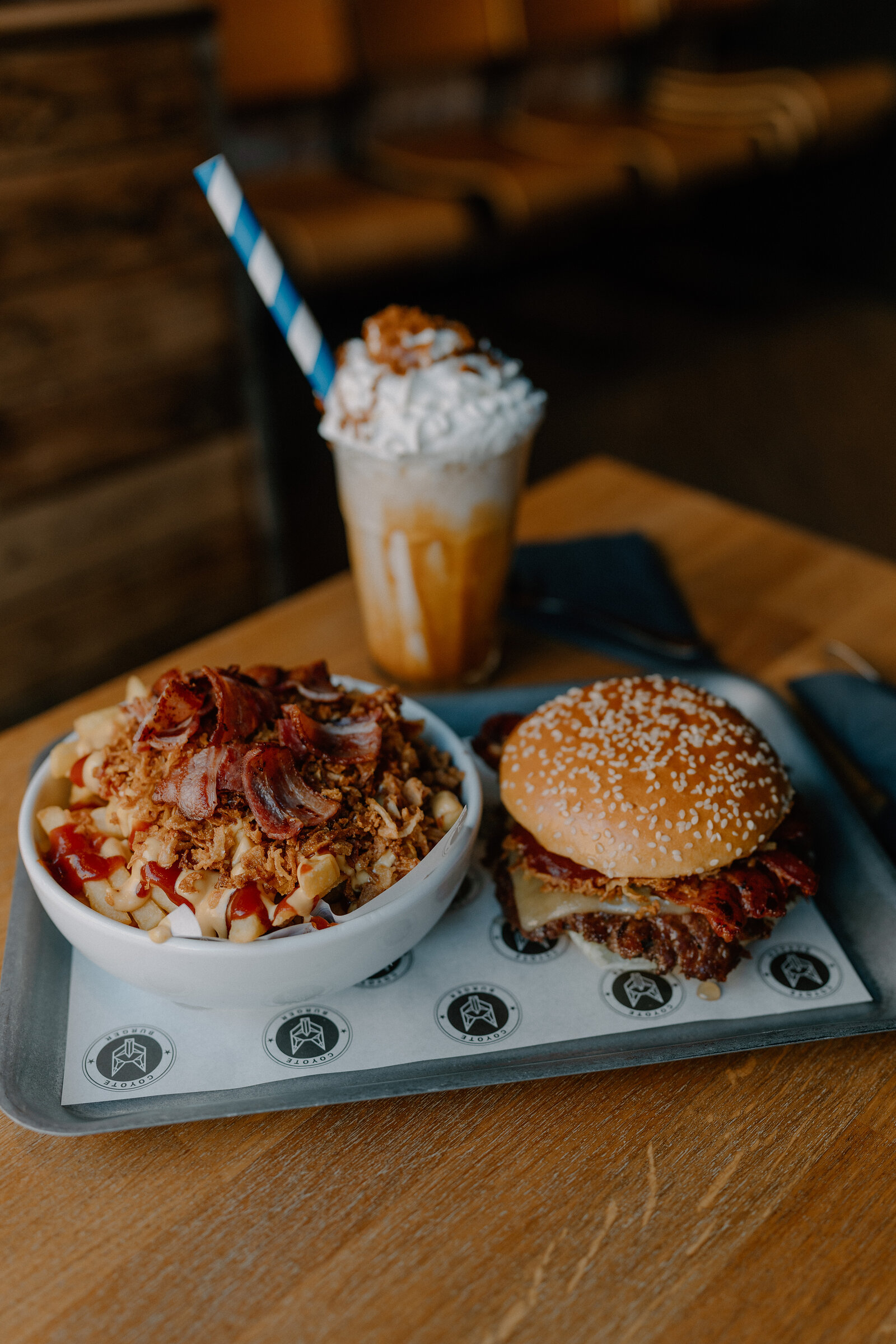 A whipped-cream topped milkshake with a striped straw stands beside a bowl of loaded fries and a sesame burger on a tray.