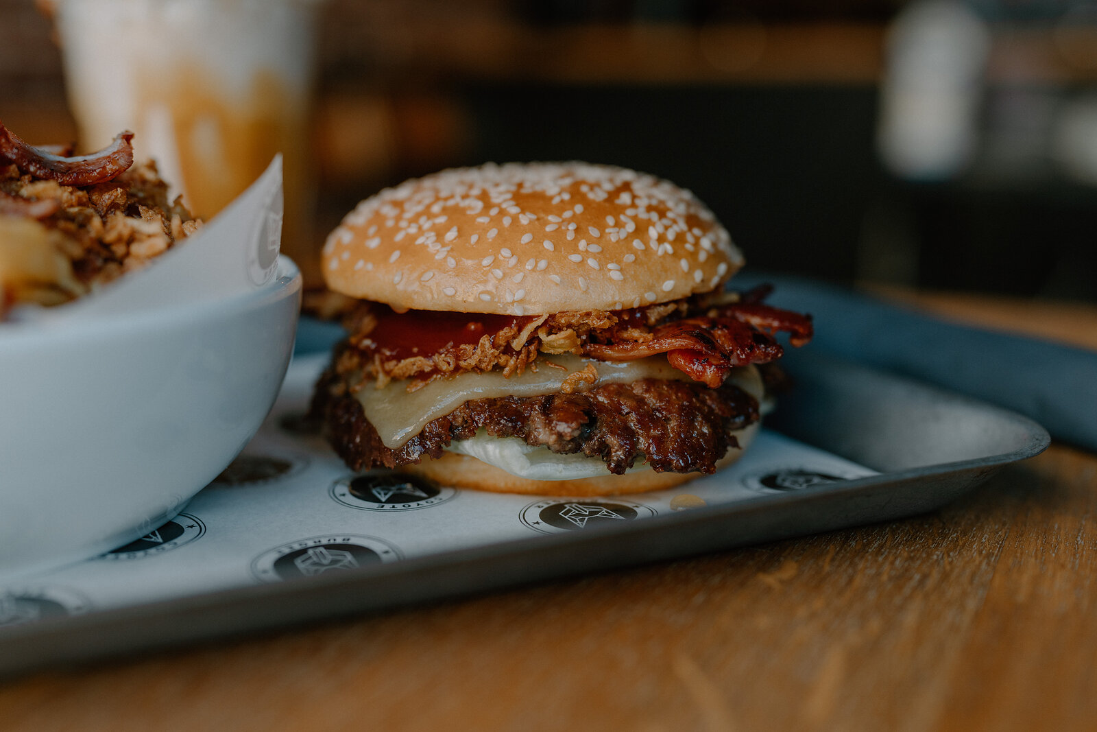 A sesame bun burger stacked with bacon, cheese and crispy onions sits on a tray beside a bowl of fries and a drink.