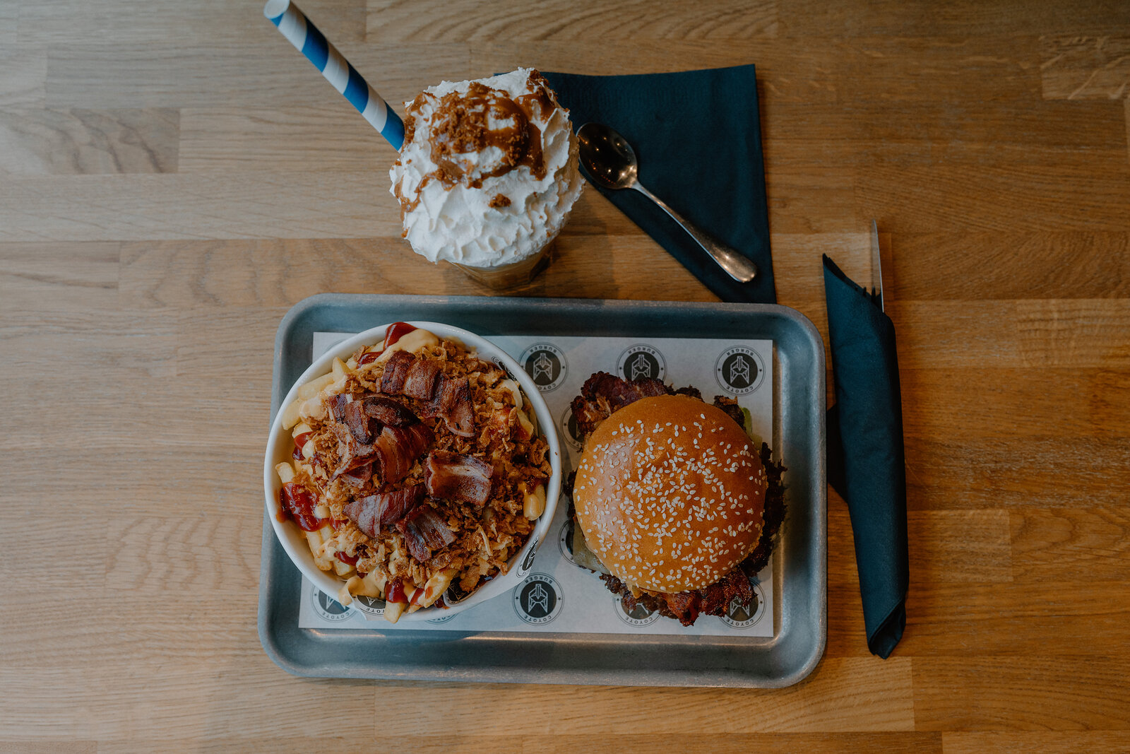 A whipped-cream topped milkshake with a striped straw stands beside a bowl of loaded fries and a sesame burger on a tray.