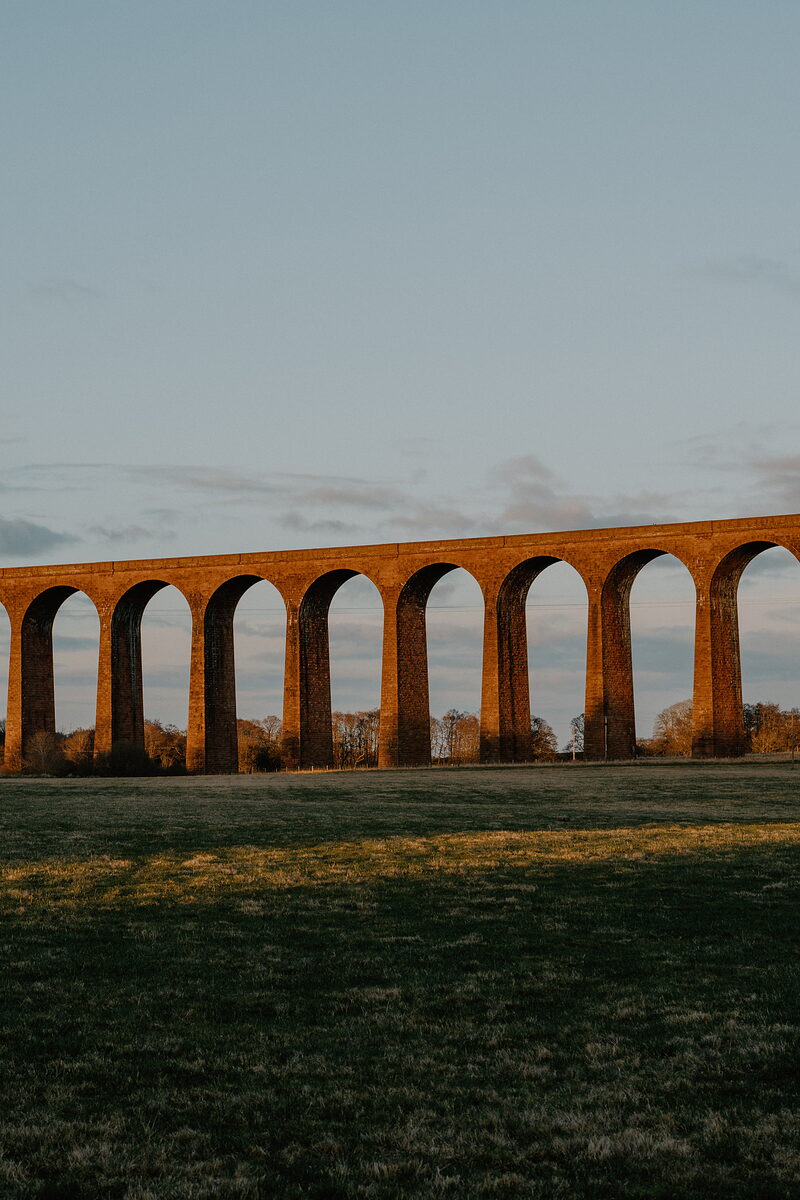 A tall brick railway viaduct with repeating arches catches warm evening light above an open field.