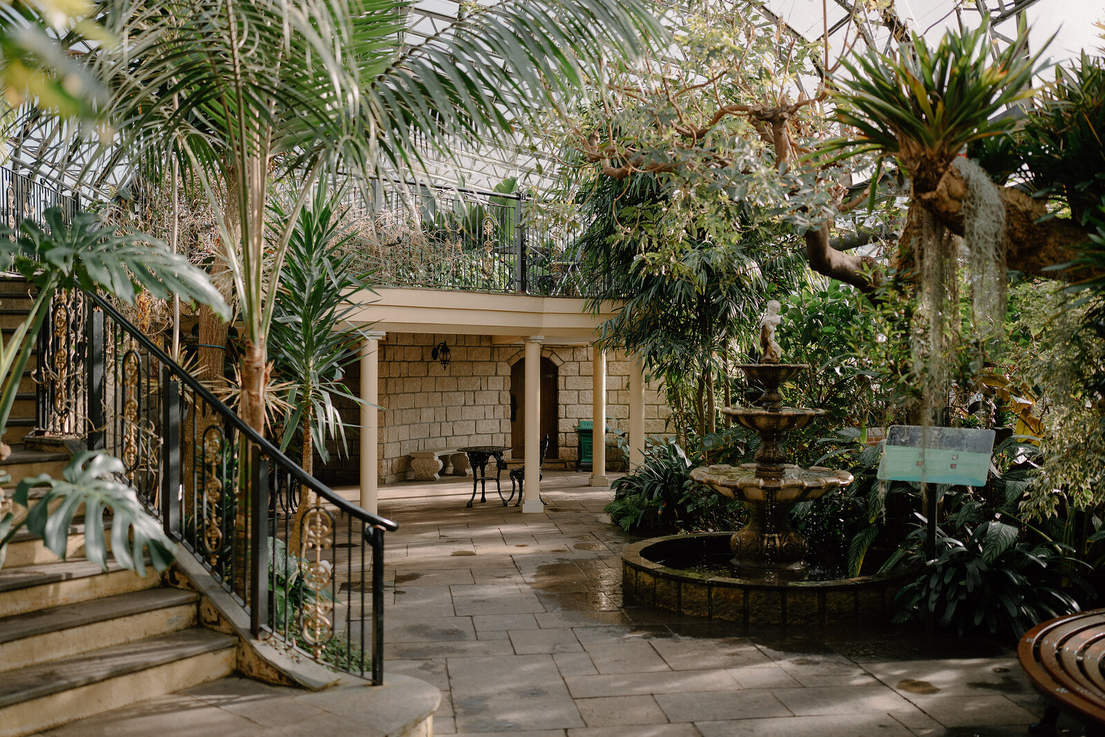 A stone fountain trickles water in a paved courtyard surrounded by palms and tropical planting, an iron staircase leading to an upper gallery.