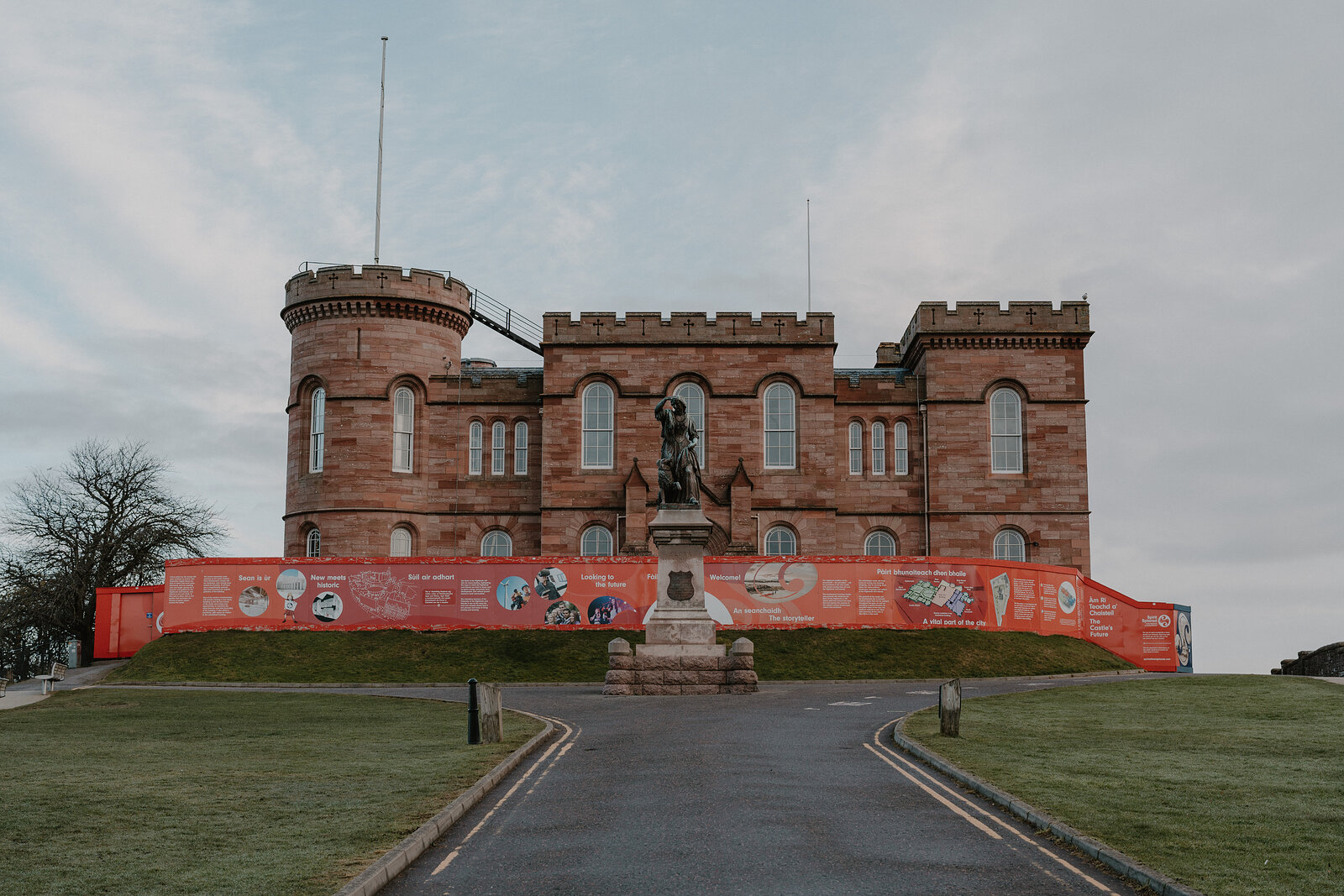 A crenellated sandstone castle with corner turrets stands on a wooded hill above a river, a statue and red hoarding visible at its base.