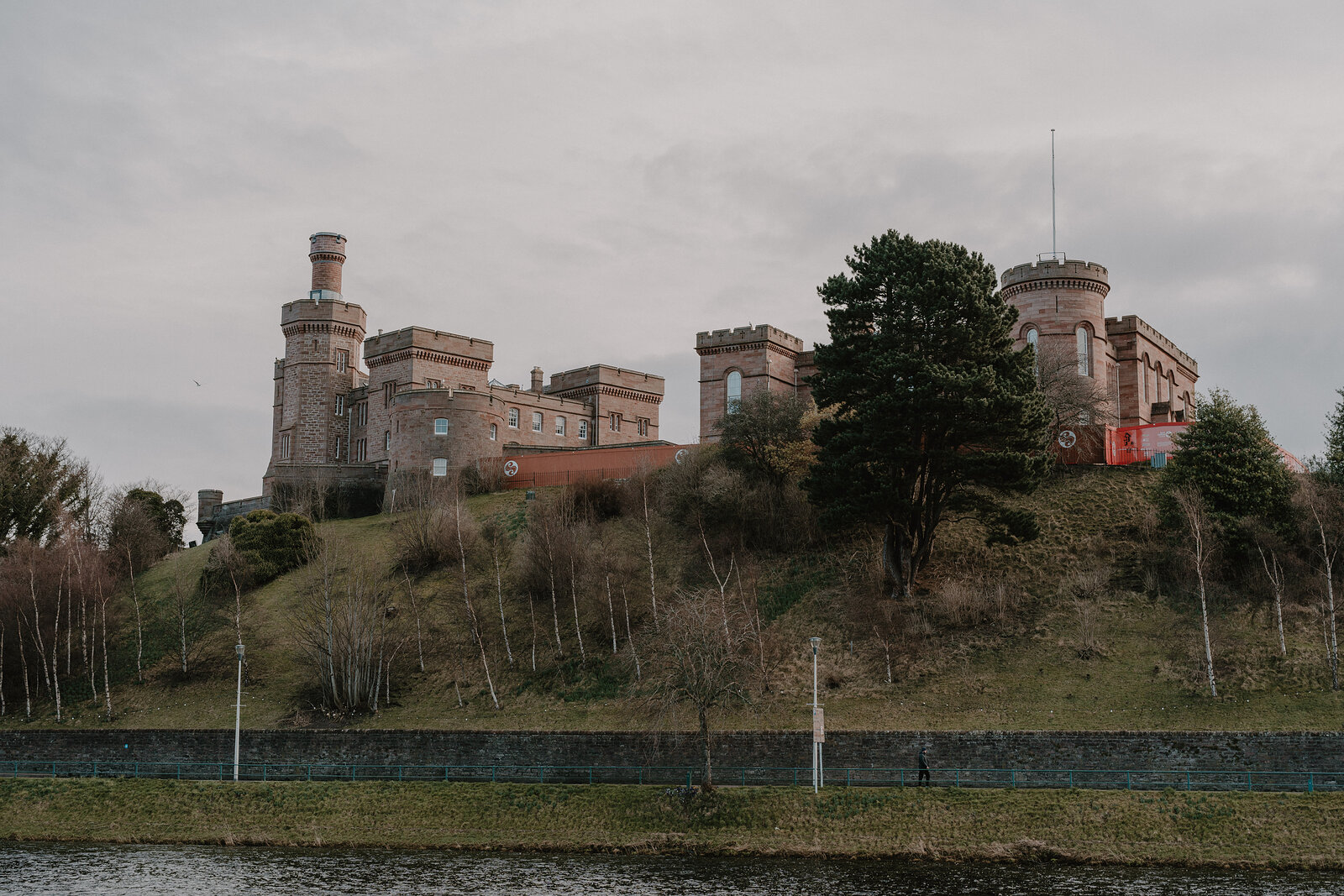A crenellated sandstone castle with corner turrets stands on a wooded hill above a river, seen from across the water.
