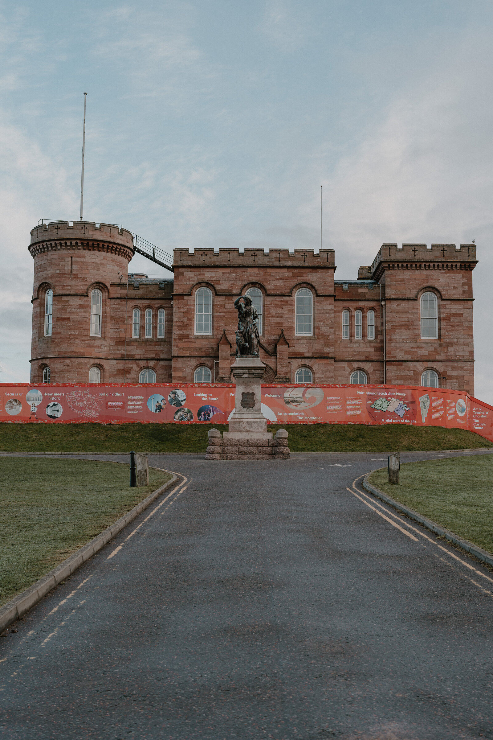 A crenellated sandstone castle with corner turrets stands behind a bronze statue on a plinth and a banner strung along the lawn.