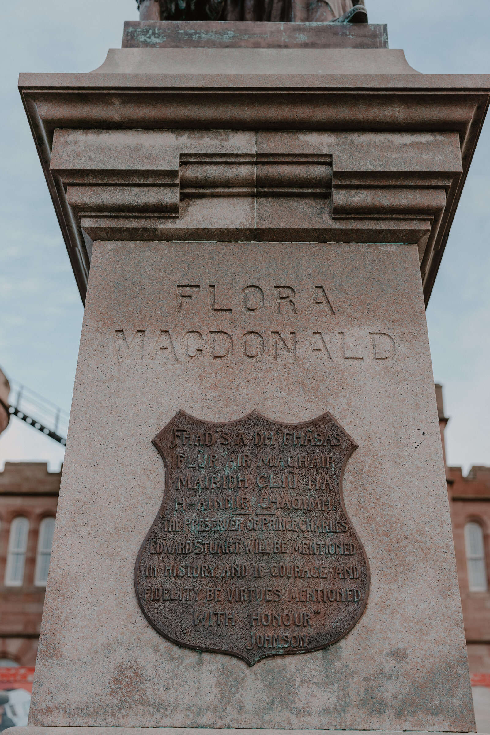 A stone plinth inscribed 'Flora MacDonald' carries a Gaelic verse and tribute beneath a shield emblem, a crenellated building visible behind.