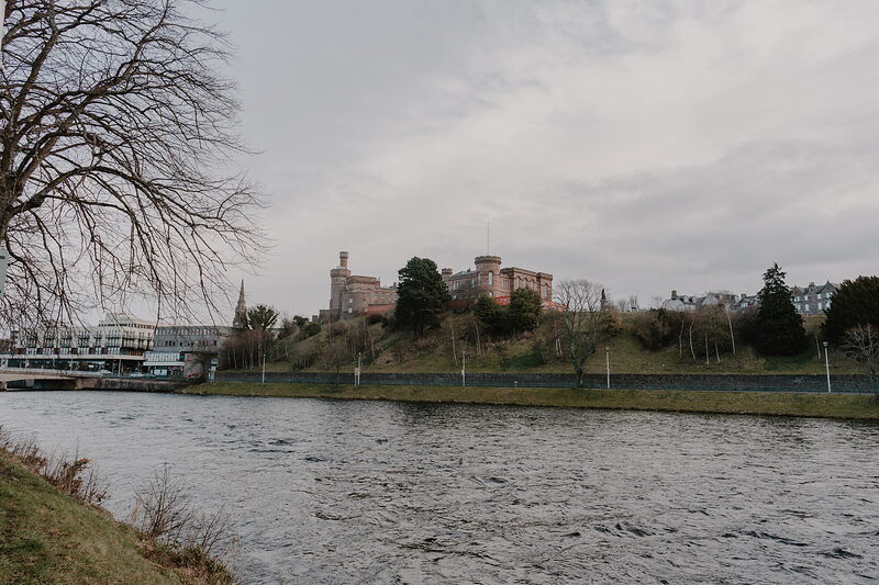 A crenellated sandstone castle stands on a wooded hill above a river, bare trees lining the bank in the foreground.