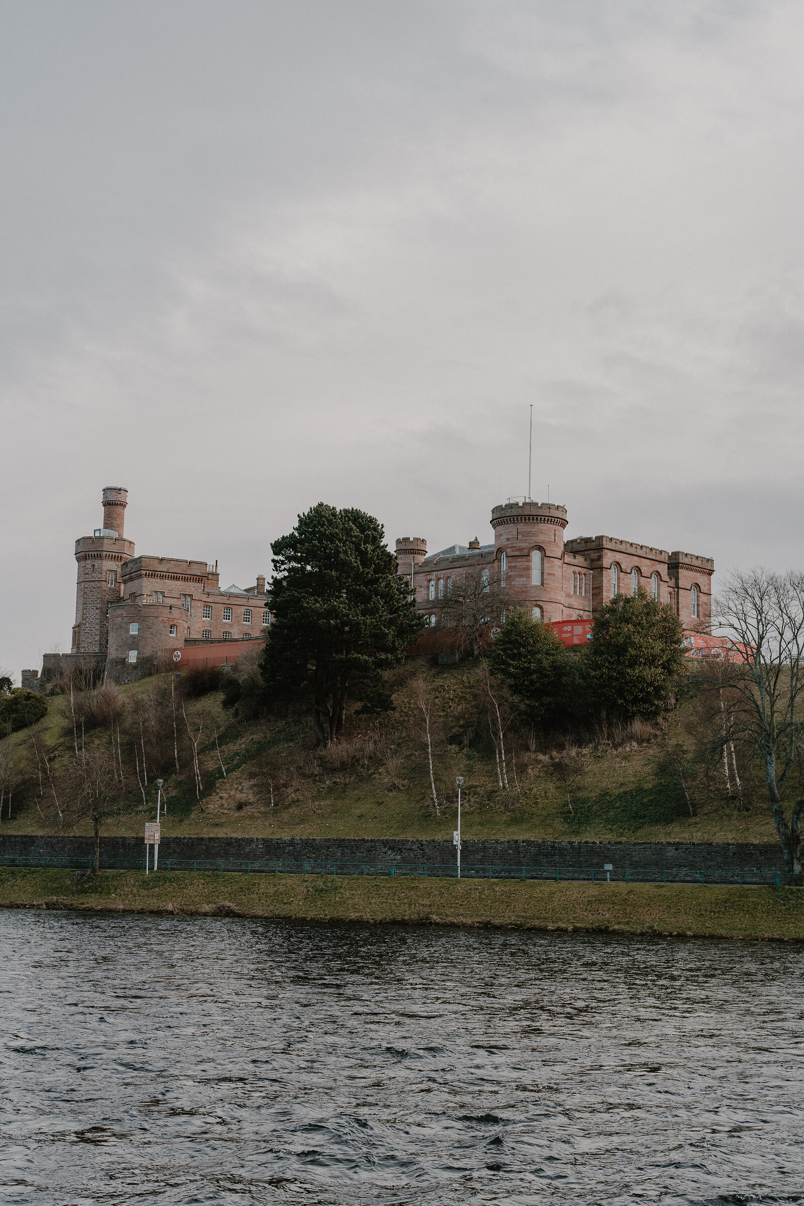 A crenellated sandstone castle with corner turrets stands on a wooded hill above a river beneath an overcast sky.