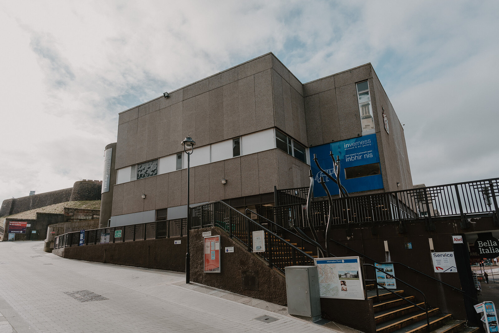 A concrete museum building with an angled staircase and a blue banner reading 'New Fib' stands on a street corner, information panels mounted beside the entrance.
