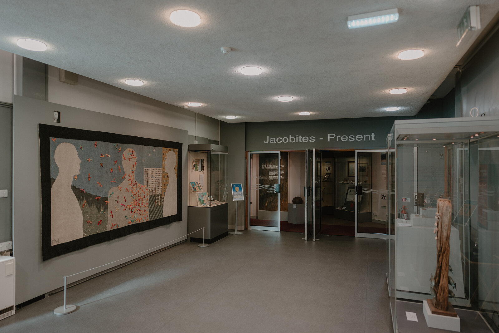 A museum corridor displays a large tapestry beside a sign reading 'Jacobites – Present', glass cases and information panels lining the hallway.