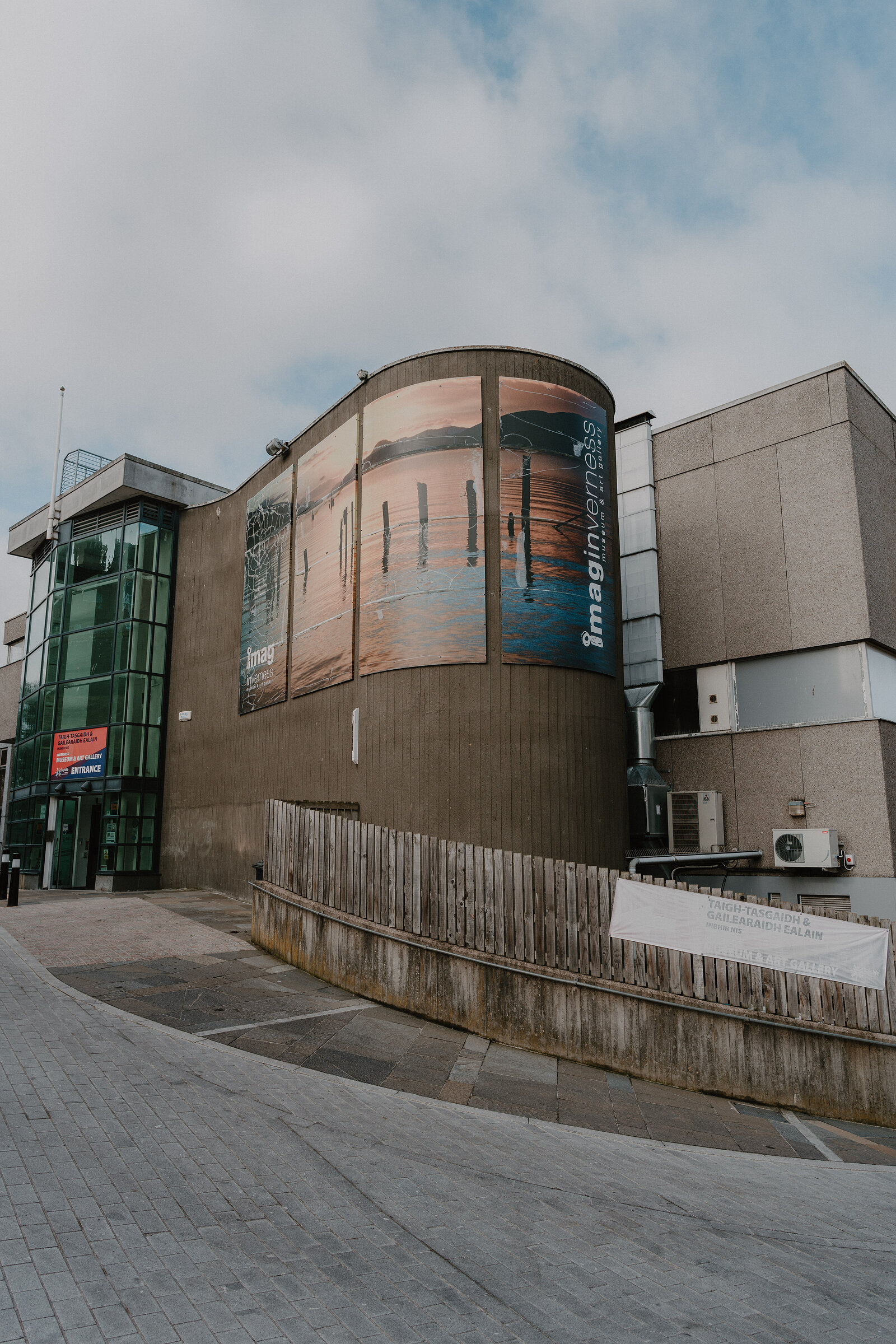 A curved concrete museum building displays large photographic banners beside a glazed entrance tower, a hoarding and parked cars nearby.