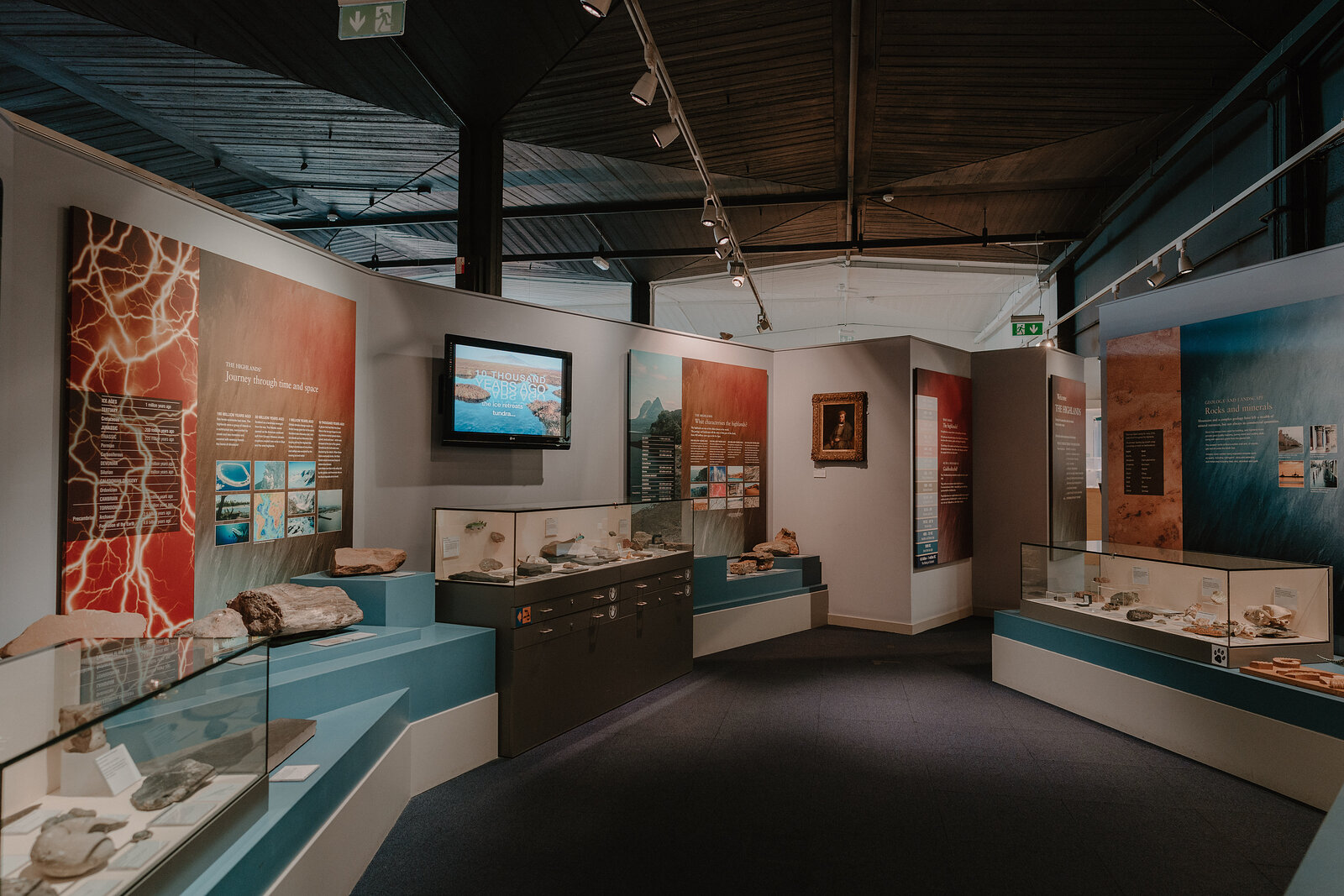 A museum gallery displays archaeological finds and information panels around a central video screen, glass cases lining both walls.