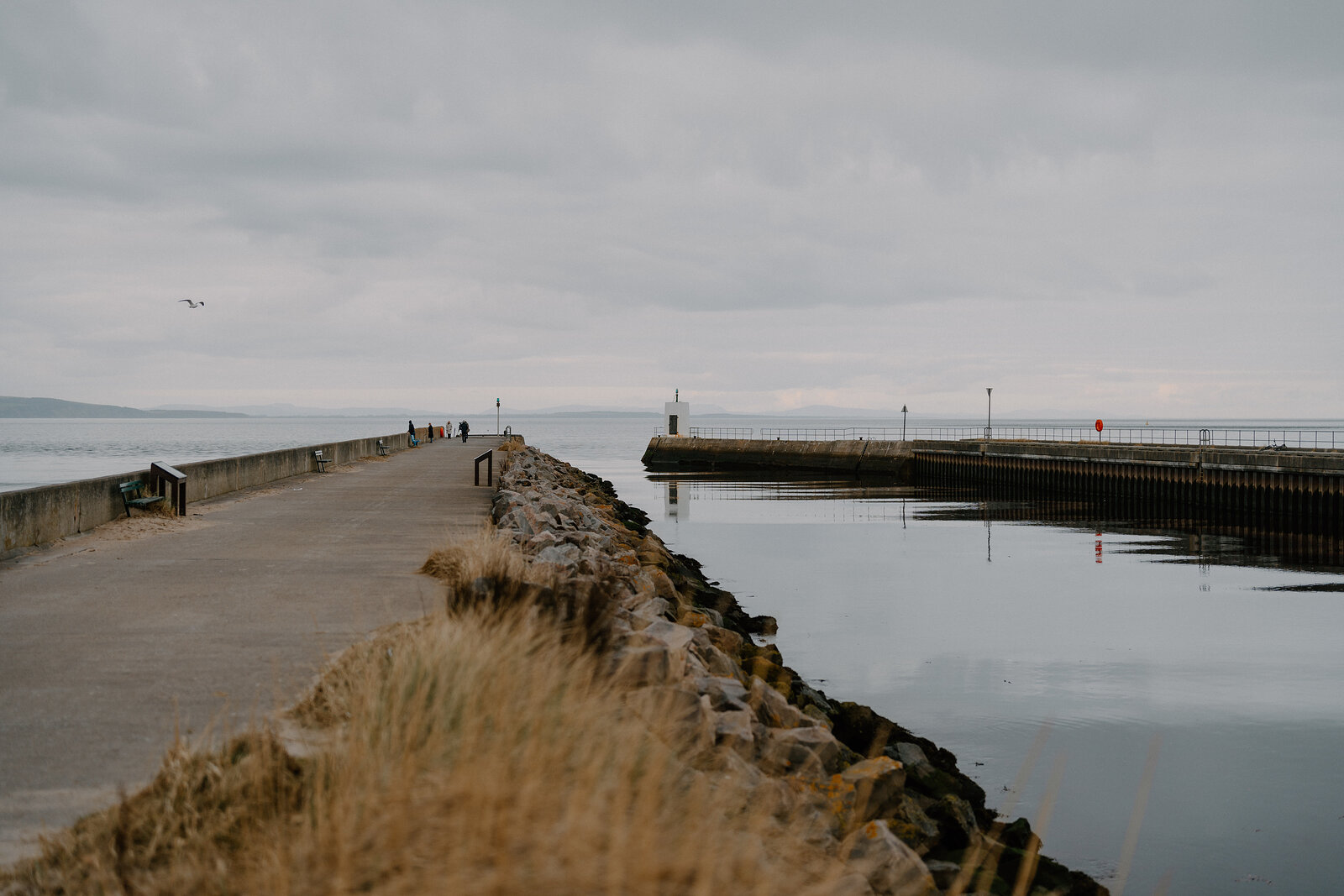 A stone breakwater curves around a calm harbour basin beside a rocky pier, people walking along the path in the distance.