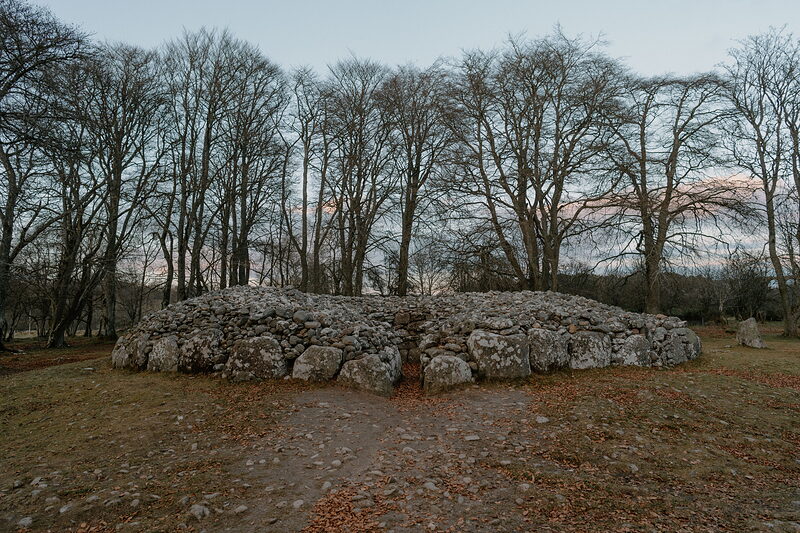 A large circular chambered cairn built from stacked stones sits among bare winter trees at dusk.