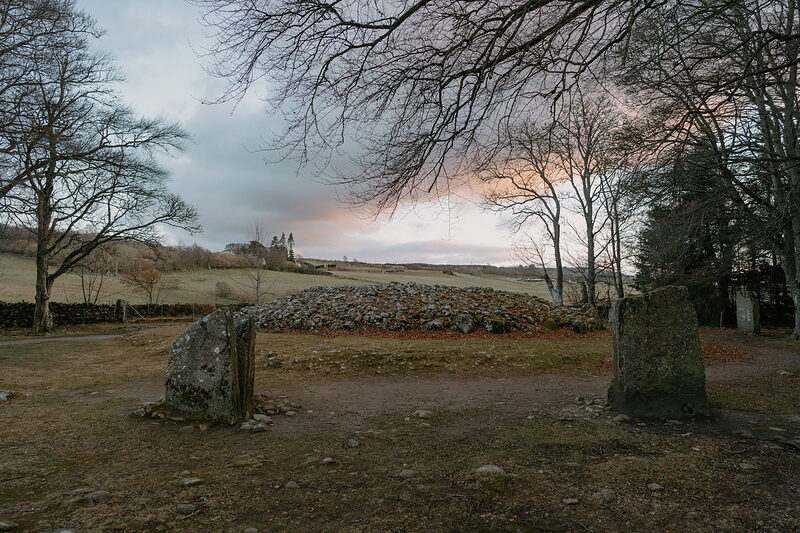 A large circular chambered cairn stands between two standing stones, bare winter trees framing a pink and grey dusk sky.