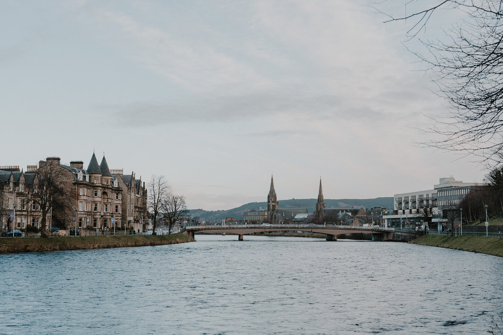 A river flows past sandstone buildings toward a stone bridge, twin church spires rising on the far bank beneath a pale sky.