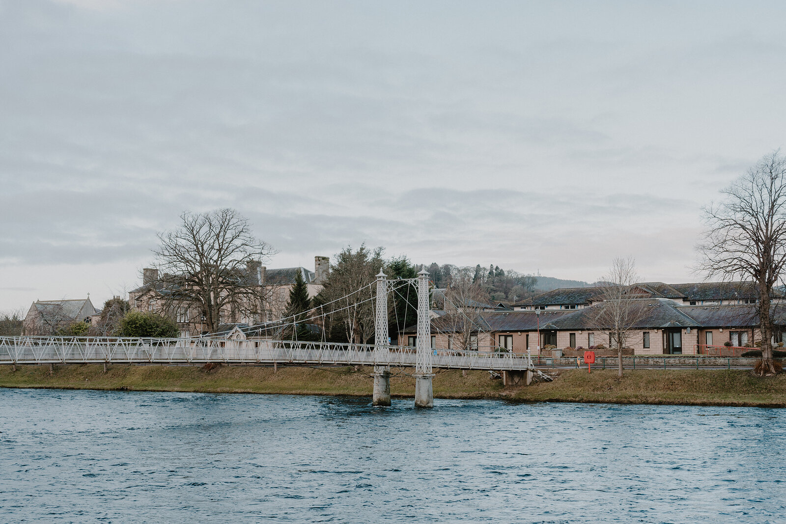 A white cast-iron suspension footbridge crosses a calm river beside a row of low buildings, bare trees framing the view.